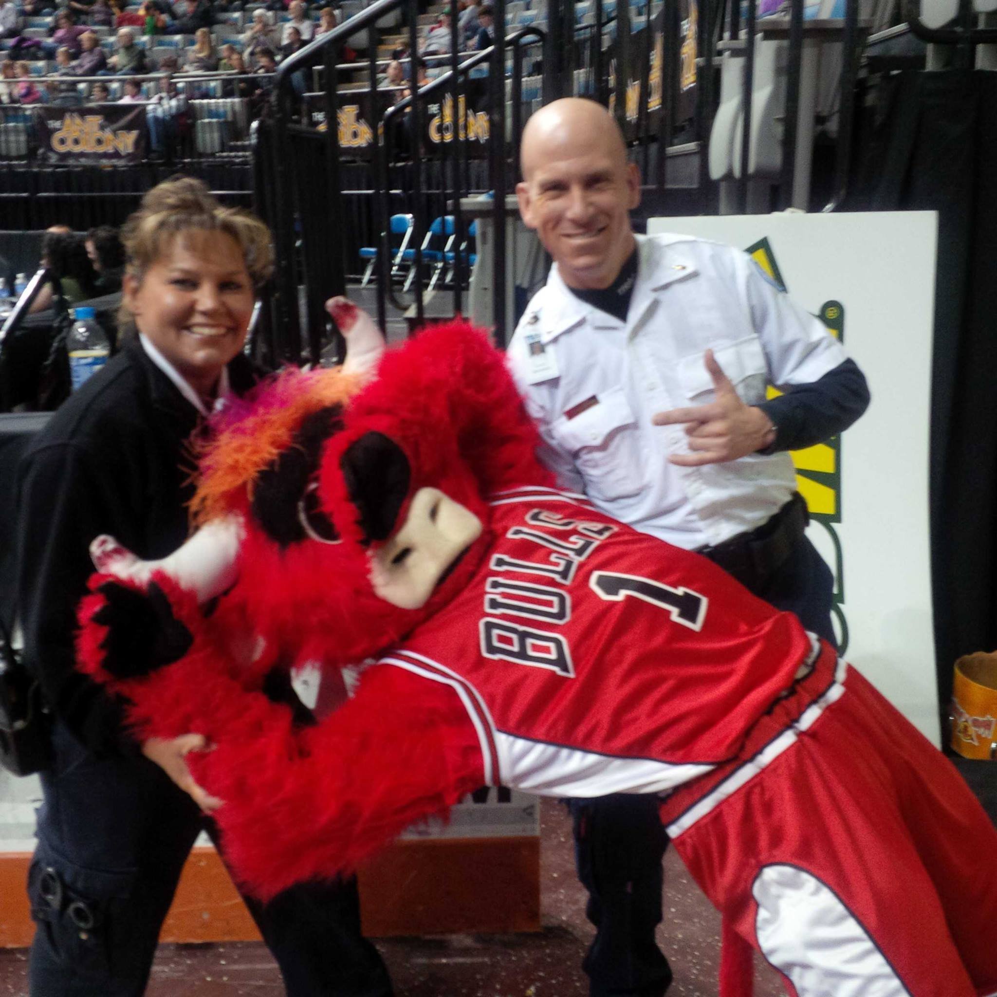 A man in a White Sox hockey jersey and Monica Kelsey in formal attire are sitting next to a person dressed in a red, black, and yellow animal costume, with a background of stadium seating filled with spectators.