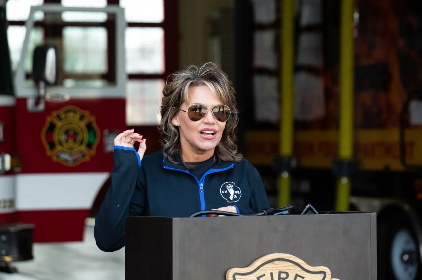 Monica Kelsey with wavy hair, wearing sunglasses and a navy jacket with a firefighter emblem, standing at a podium with a fire station in the background.
