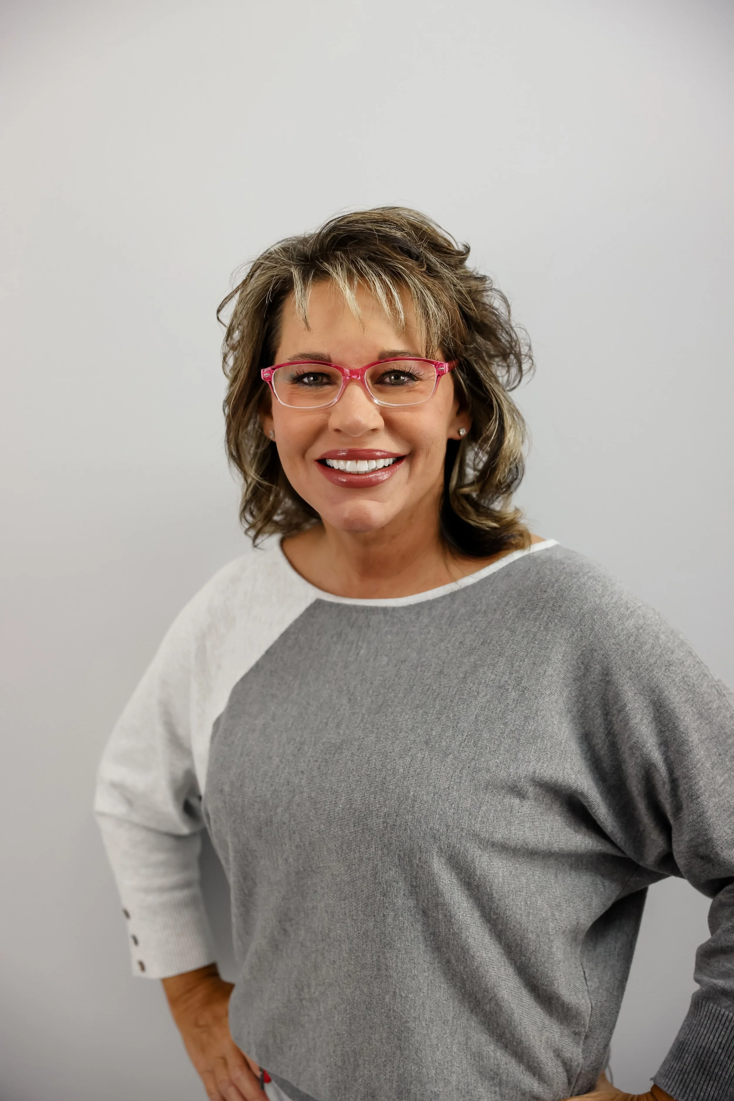 Monica Kelsey smiling, wearing pink glasses and a white and gray sweatshirt, standing against a plain gray background.