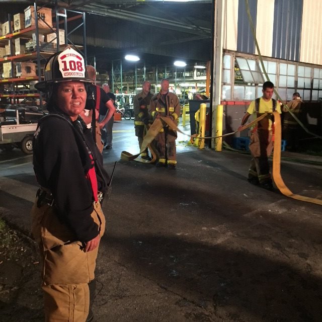 Monica Kelsey wearing a helmet and tan firefighting gear, standing outside at night with firefighters in the background, some holding fire hoses.