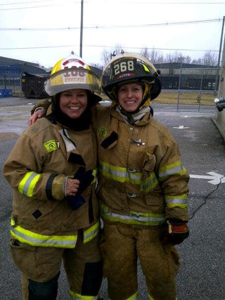Two firefighters in full gear smiling and standing side by side in a parking lot.