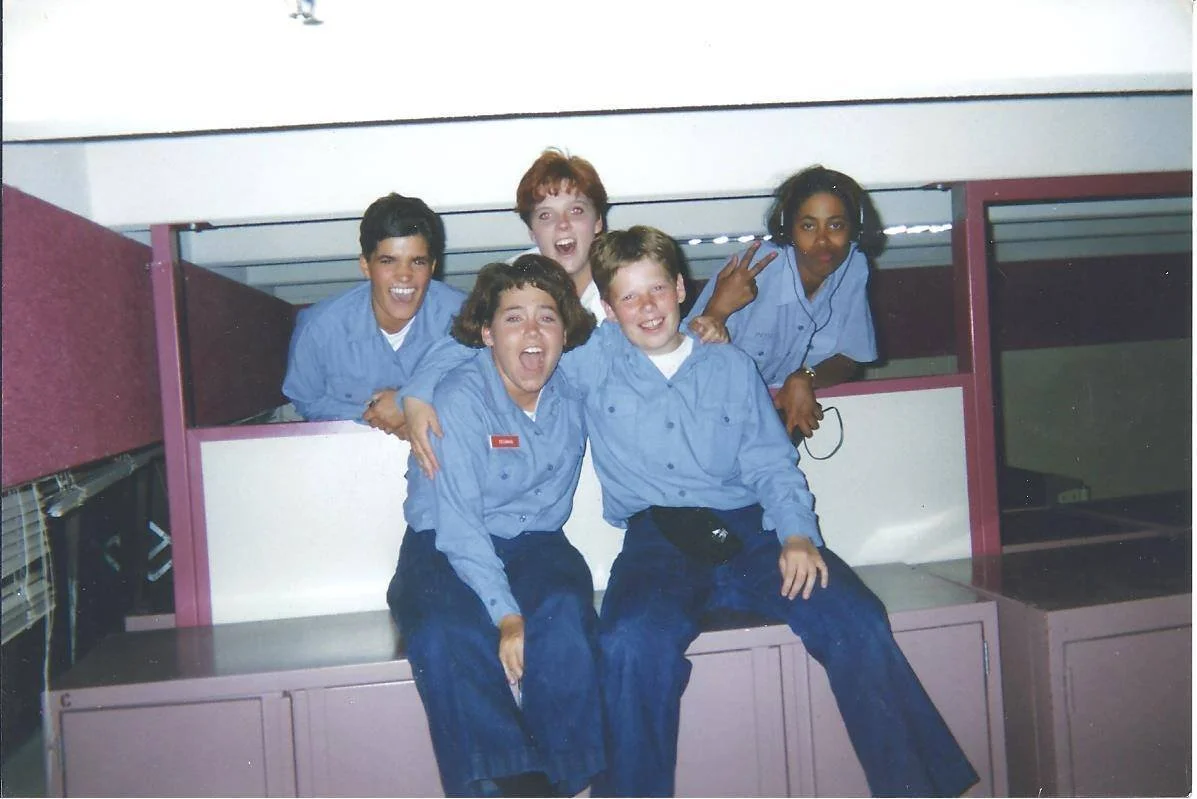 Five teenagers in blue uniform shirts and dark pants posing and smiling inside a school cubicle area.