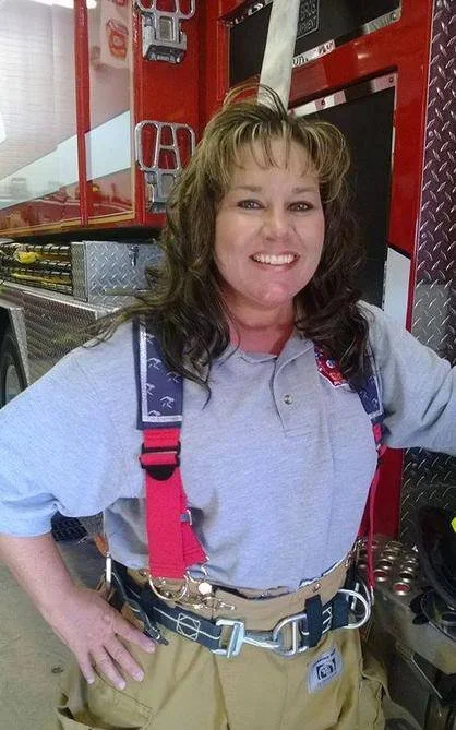 Monica Kelsey in firefighting gear inside a fire station, smiling with her hand on her hip, surrounded by firefighting equipment and a red fire truck in the background.