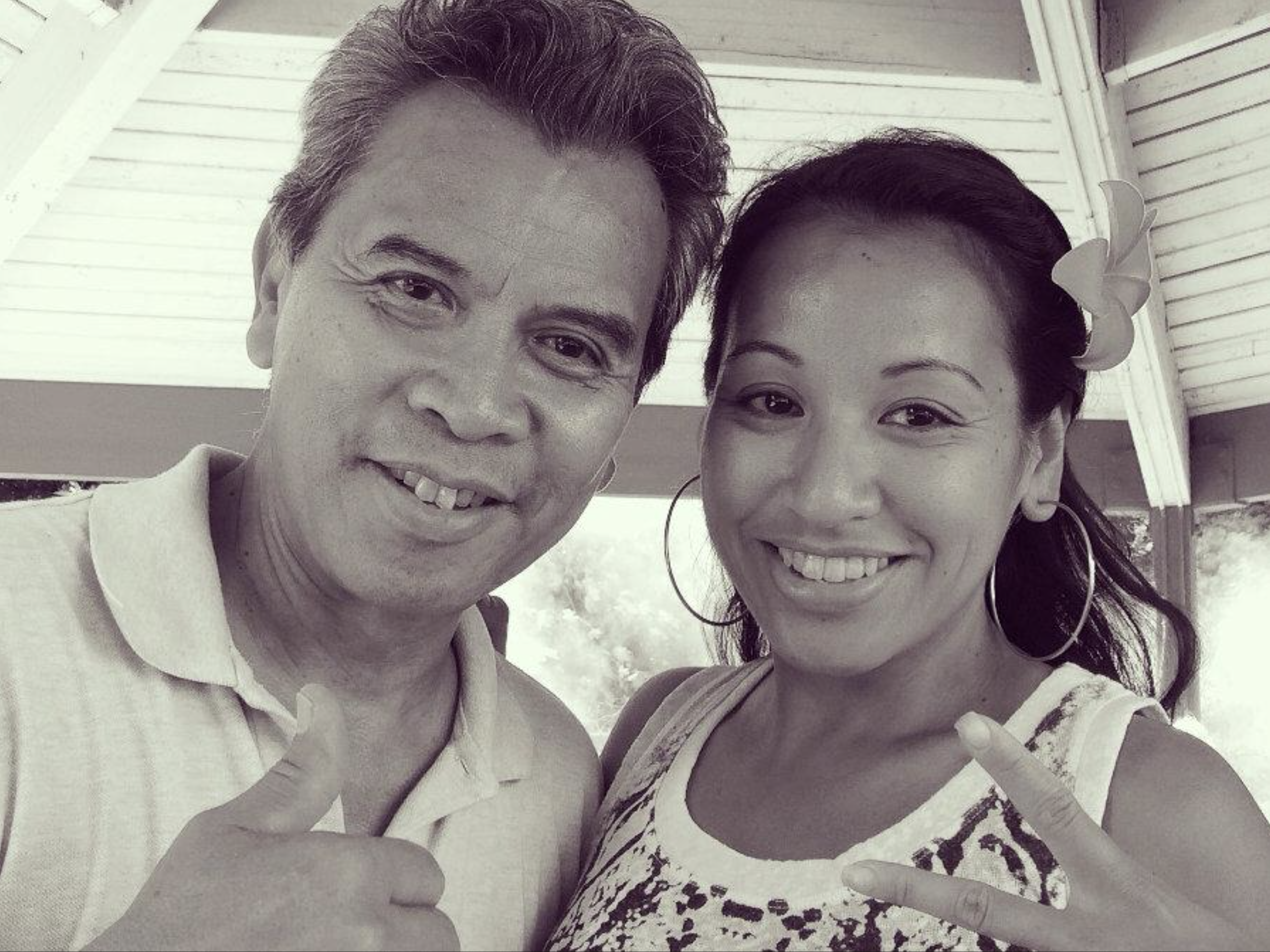 A black-and-white photo of Marisa and her father, Benny taking a selfie outdoors. Both are smiling, dressed casually, and there are trees and a building with a sloped roof in the background.