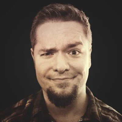 A young man with short brown hair, a beard, and a mustache, wearing a plaid shirt, looking at the camera with a playful expression and one eye slightly squinted, against a dark background.