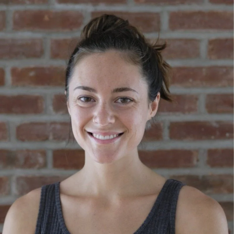 A woman with light skin and dark brown hair tied in a messy bun smiling in front of a brick wall.