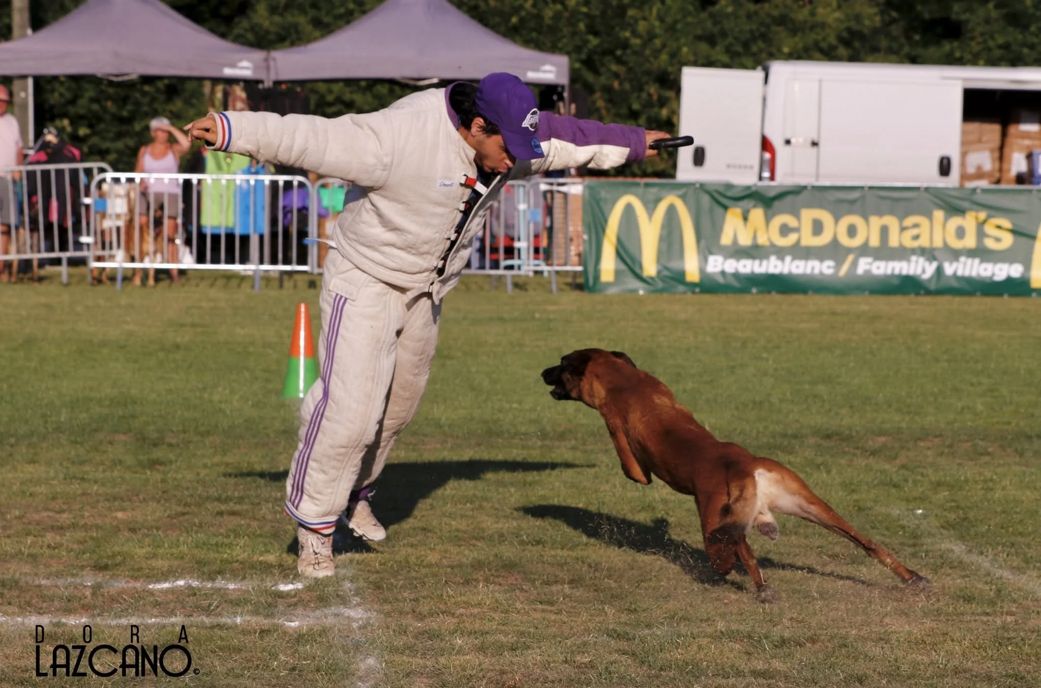 A man in a racing suit and purple cap leaning forward with arms outstretched above the back of a brown dog as they participate in a dog obedience or agility event outdoors, with spectators, tents, and a McDonald's banner in the background.