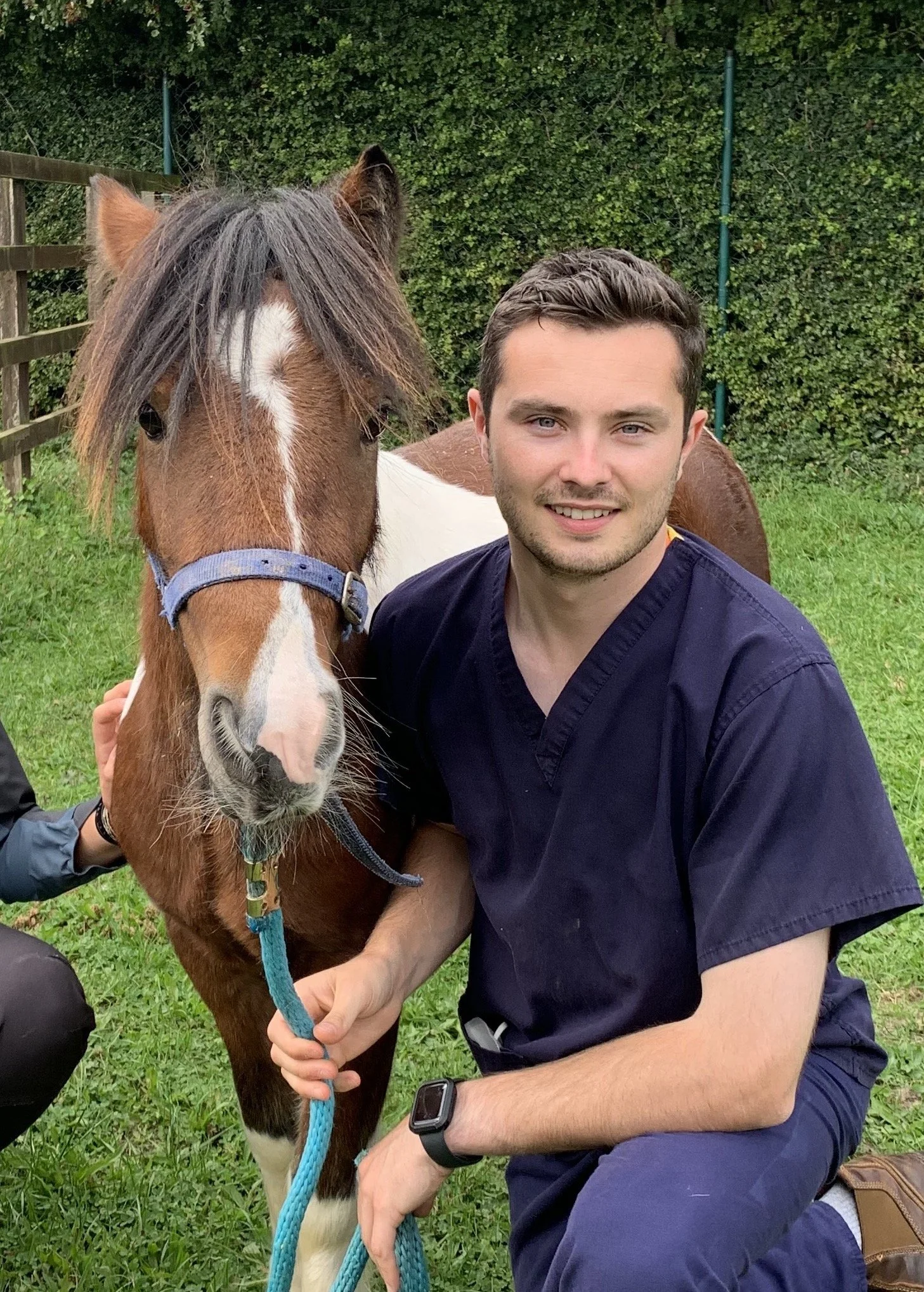 A young man with short dark hair and blue eyes, wearing a black shirt and a smartwatch, smiling while kneeling next to a brown and white horse with a blue halter, in a grassy outdoor area with a wooden fence and green bushes in the background.