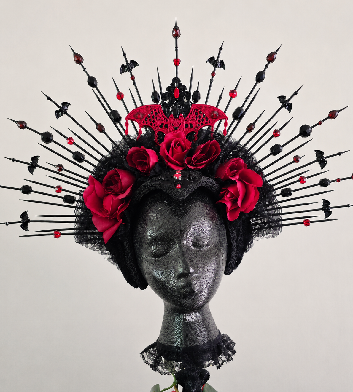 A mannequin head with metallic texture, adorned with a black lace headpiece decorated with red roses, black lace, and a red bat-shaped ornament. Black and red decorative pins radiate outward from the head.