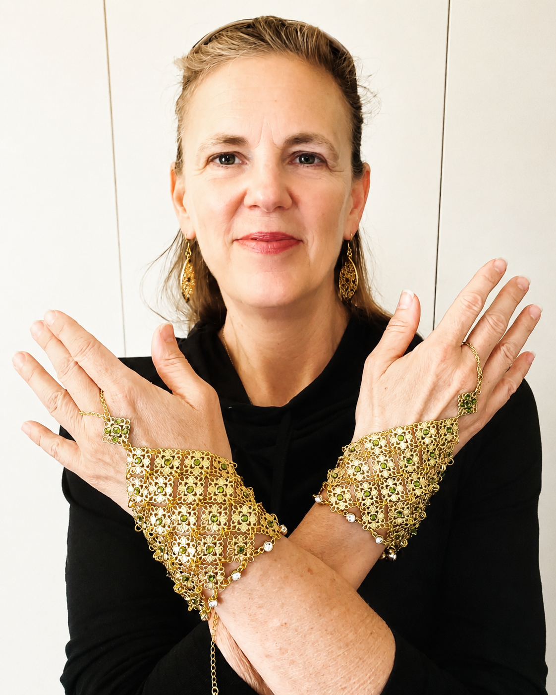A woman with blonde hair wearing gold jewelry, including earrings, bracelets, and rings, crossing her arms in front of her chest, standing against a plain background.