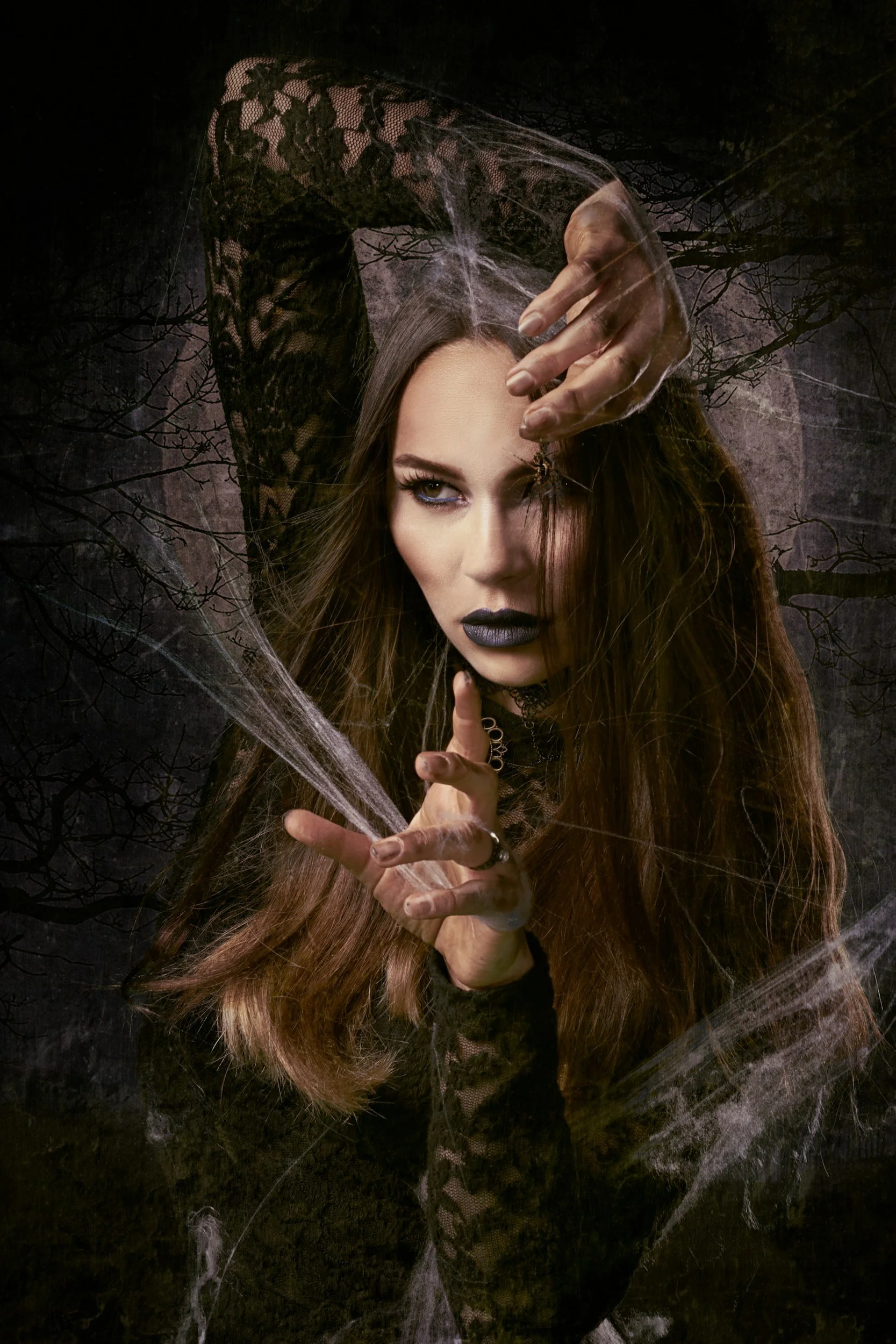 A woman with long brown hair and dark makeup, wearing a black lace top, poses amidst cobwebs in a dark outdoor setting with leafless tree branches in the background.