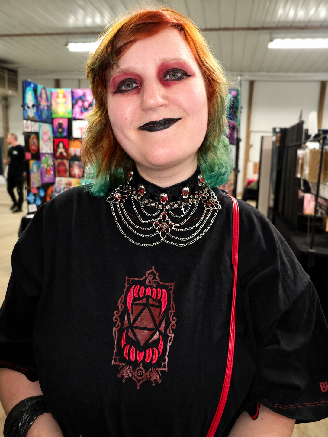 A young woman with red and green hair and gothic makeup, wearing a black gothic shirt with a red and black design, layered chains and jewelry, at an indoor art or craft market with colorful artwork in the background.