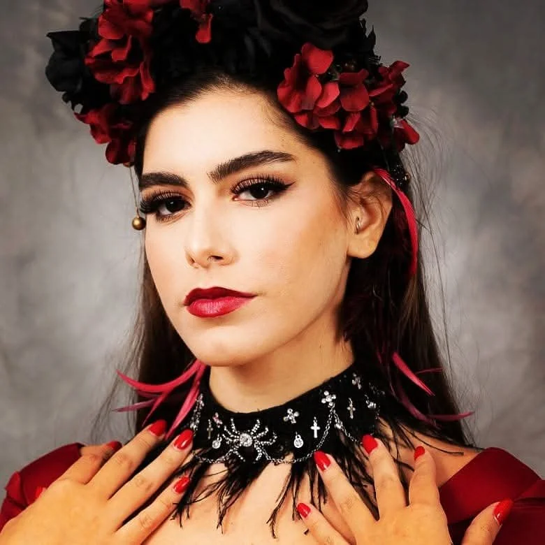 A woman with dark hair, adorned with a black and red floral headpiece, wearing makeup with red lipstick and dramatic eye makeup. She has a black embellished choker with silver jewelry motifs and matching red painted nails. The background is a neutral