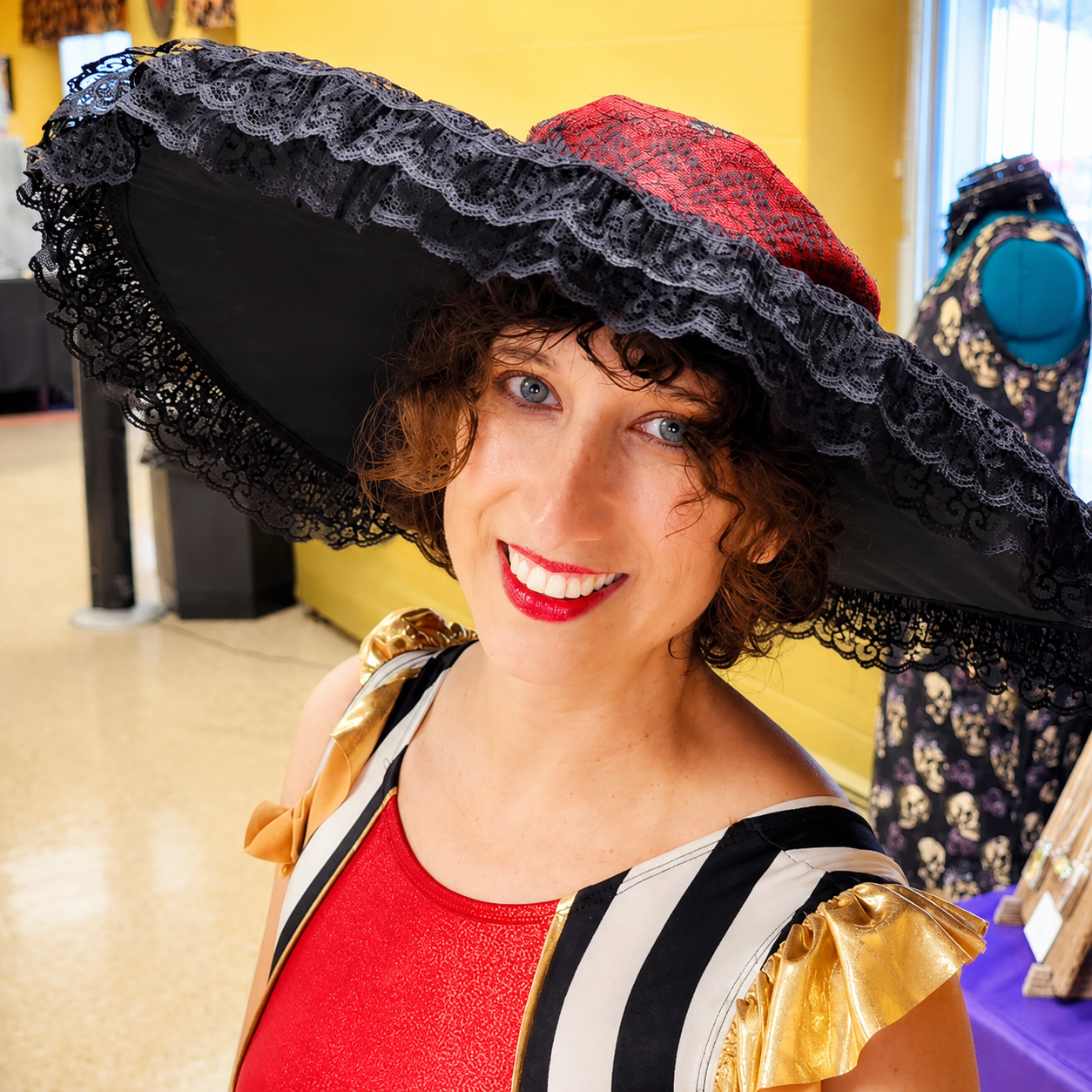 Woman smiling wearing a large black and red lace hat, red dress with gold and black striped details, and gold shoulder accents, in a room with mannequins in the background.