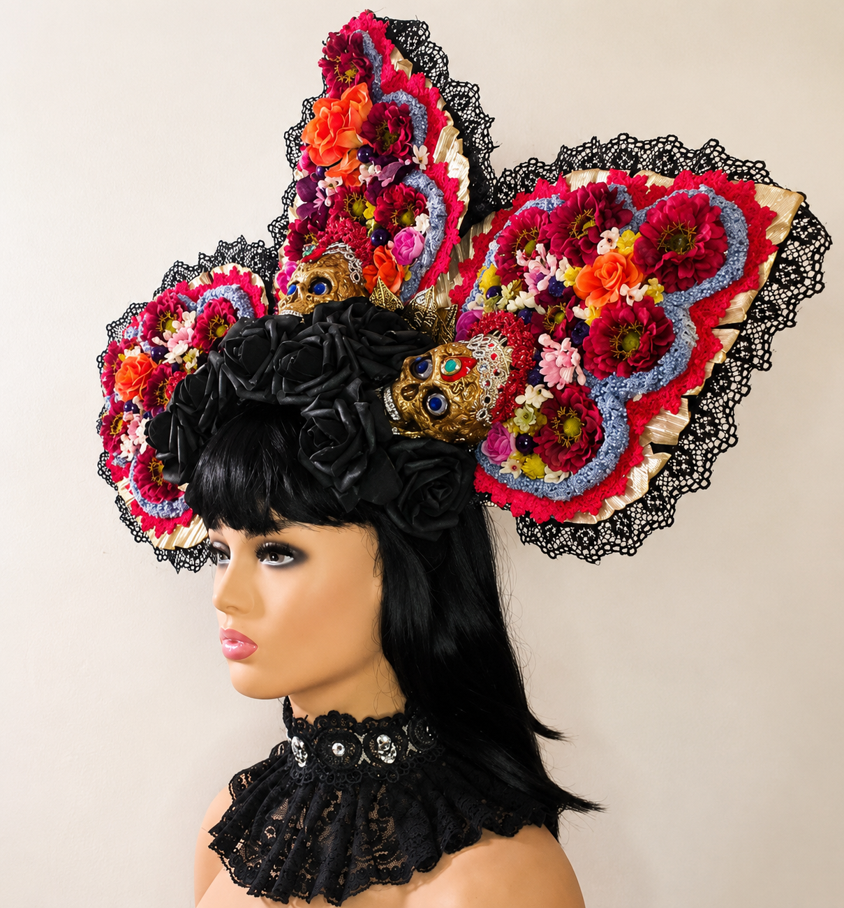 A mannequin wearing a large, colorful, floral headpiece with black lace, gold skull accents, and pink, red, orange, purple, and white flowers, dressed with a black lace choker.