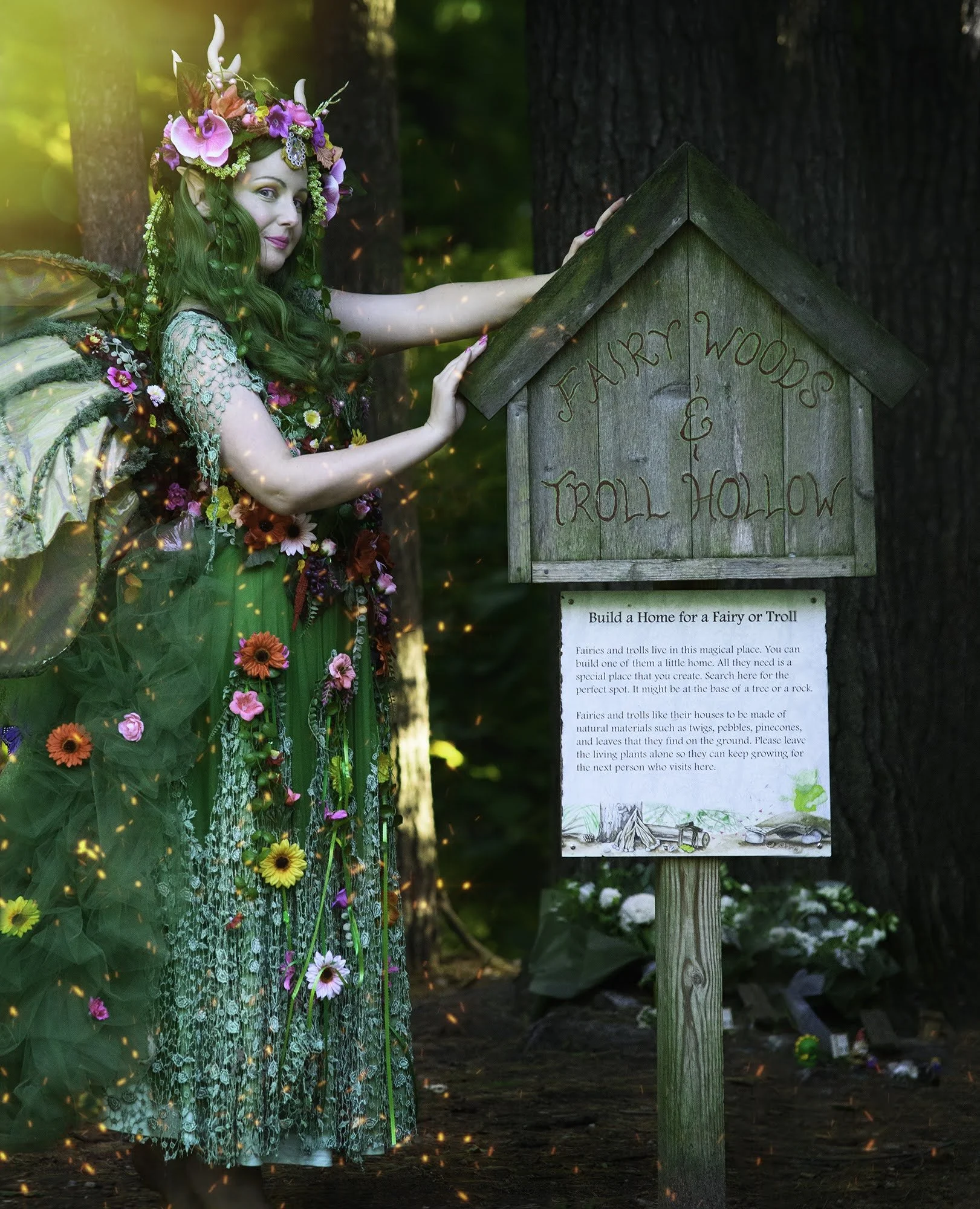 A woman dressed as a fairy with green hair, floral crown, and wing costume standing next to a wooden sign that reads 'Fairy Woods & Troll Hollow' in a wooded area.
