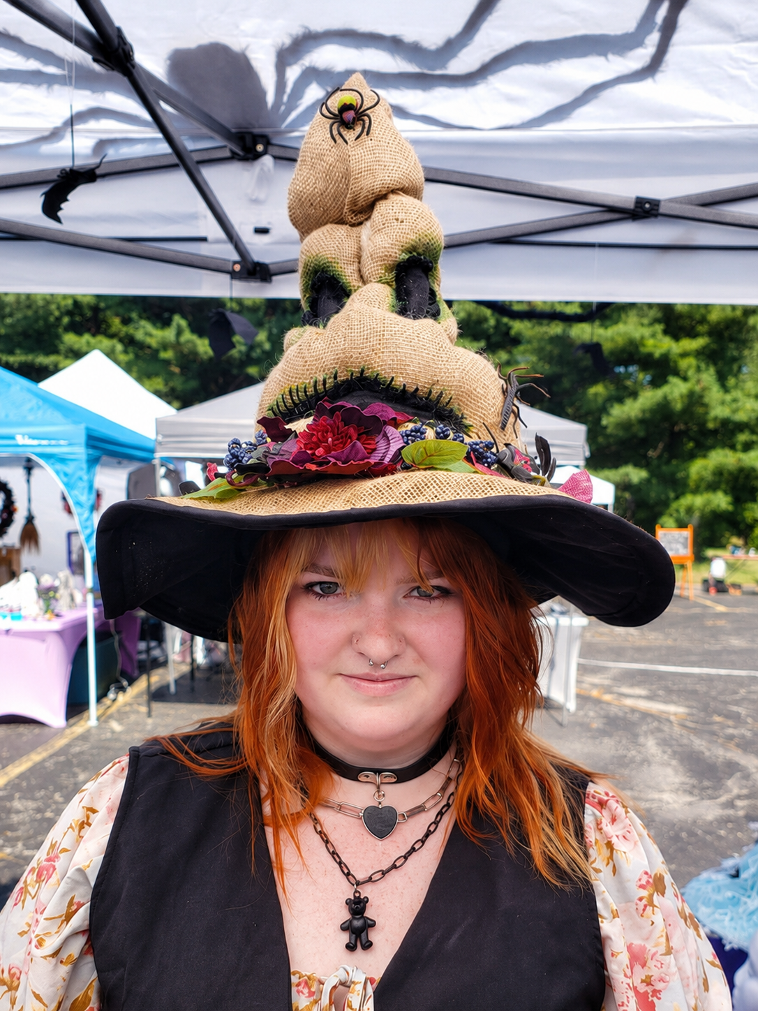 A woman with orange hair wearing a large, decorated witch hat with a spider, flowers, and berries, at an outdoor fair or market.