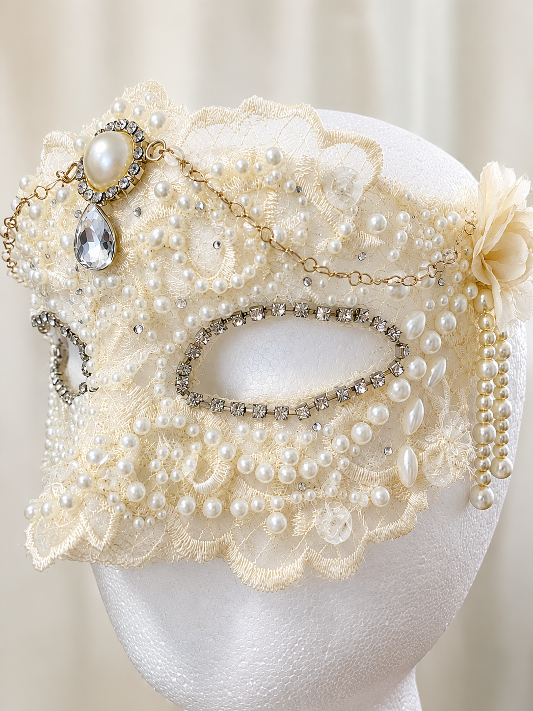 Cream-colored lace mask decorated with pearls, rhinestones, and a large crystal pendant, displayed on a white mannequin head.