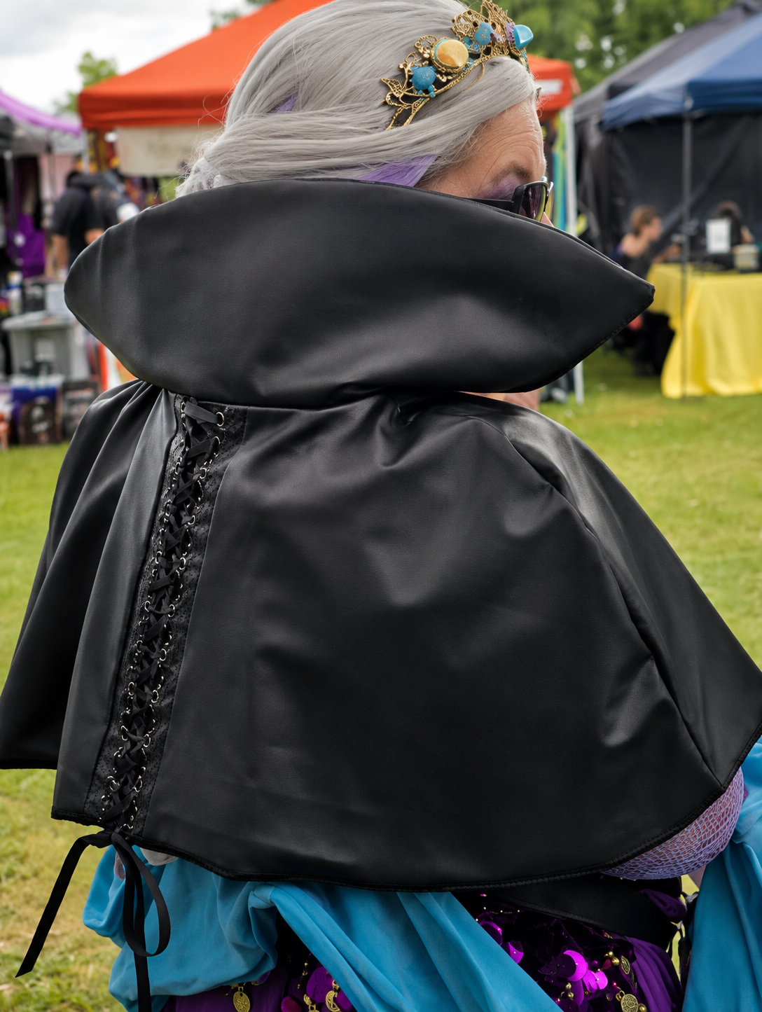 Person with long gray hair, wearing a colorful headband, sunglasses, and a dramatic black cape with lacing, at an outdoor event with tents and people in the background.