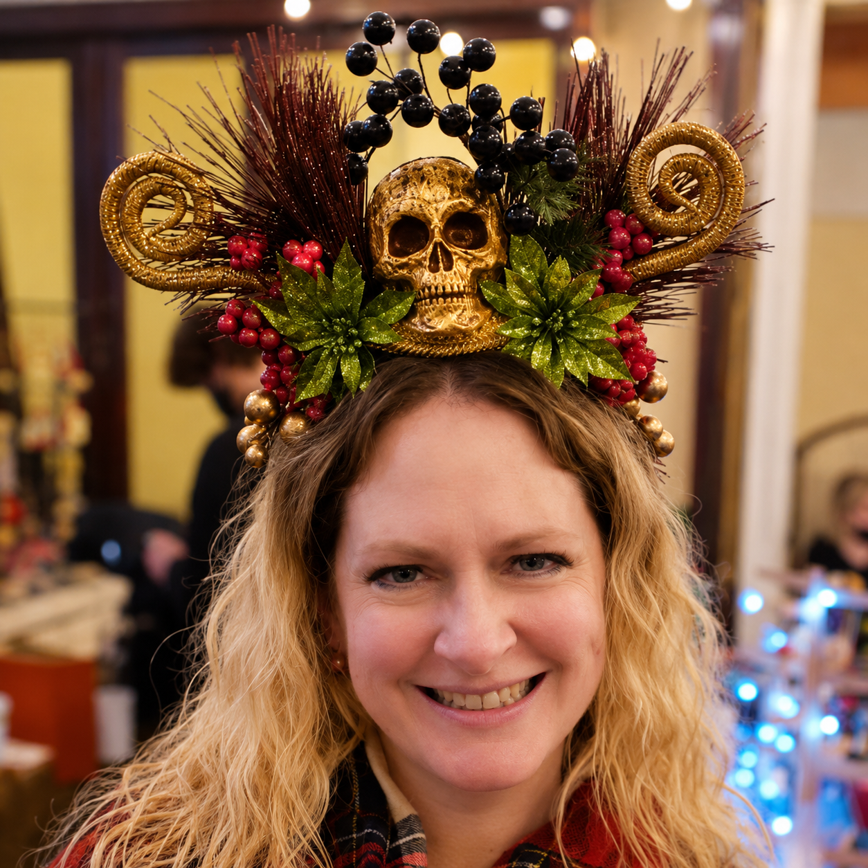 A woman with blonde hair smiling, wearing a decorative holiday hat with a gold skull, black berries, green leaves, and gold accents.