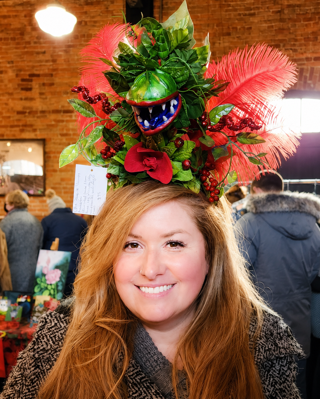 Woman with long red hair wearing a large, colorful, Christmas-themed headpiece with a plant, red feathers, red berries, and a smiling, open-mouthed toy plant monster.