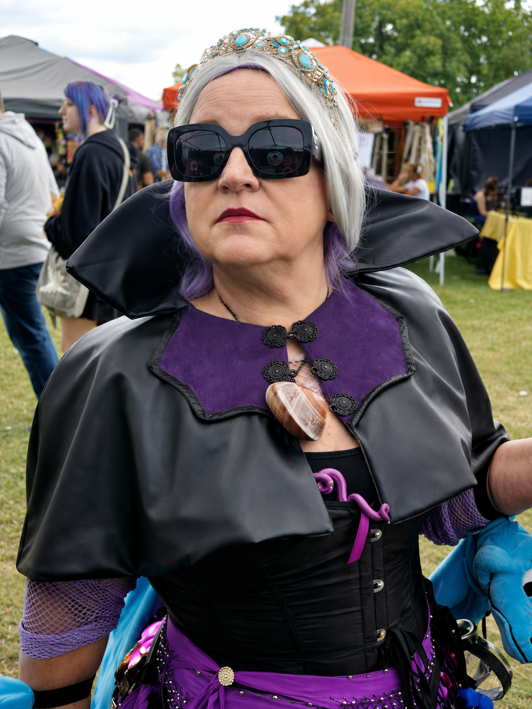Woman dressed in a black and purple costume with a large stone necklace, wearing black sunglasses, and a decorative gold and blue headpiece, at an outdoor event with tents and people in the background.