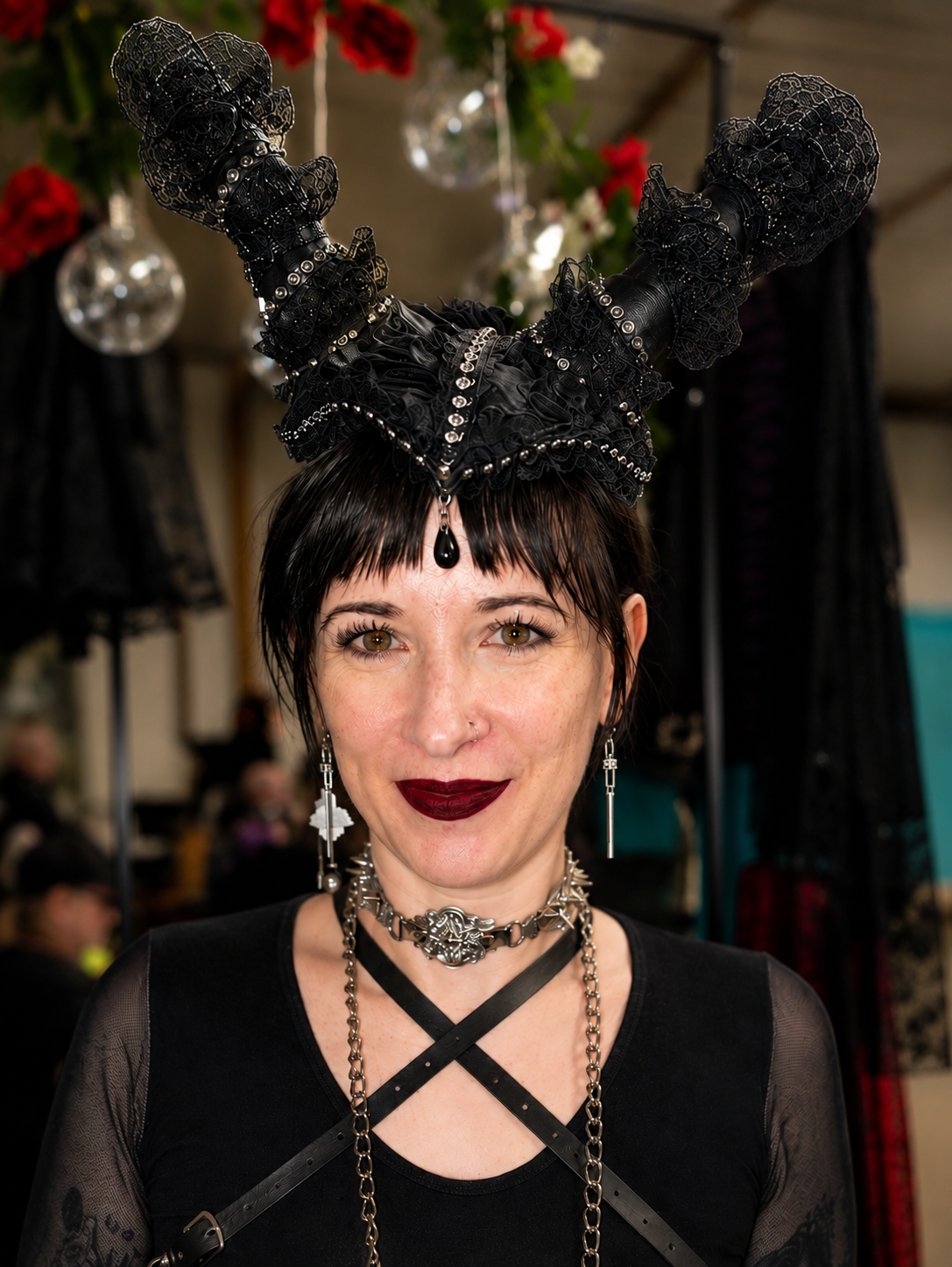 Woman with dark hair and dark lipstick wearing jewelry and a headpiece with large black lace horns, in a setting with decorative red and white flowers and hanging glass ornaments.