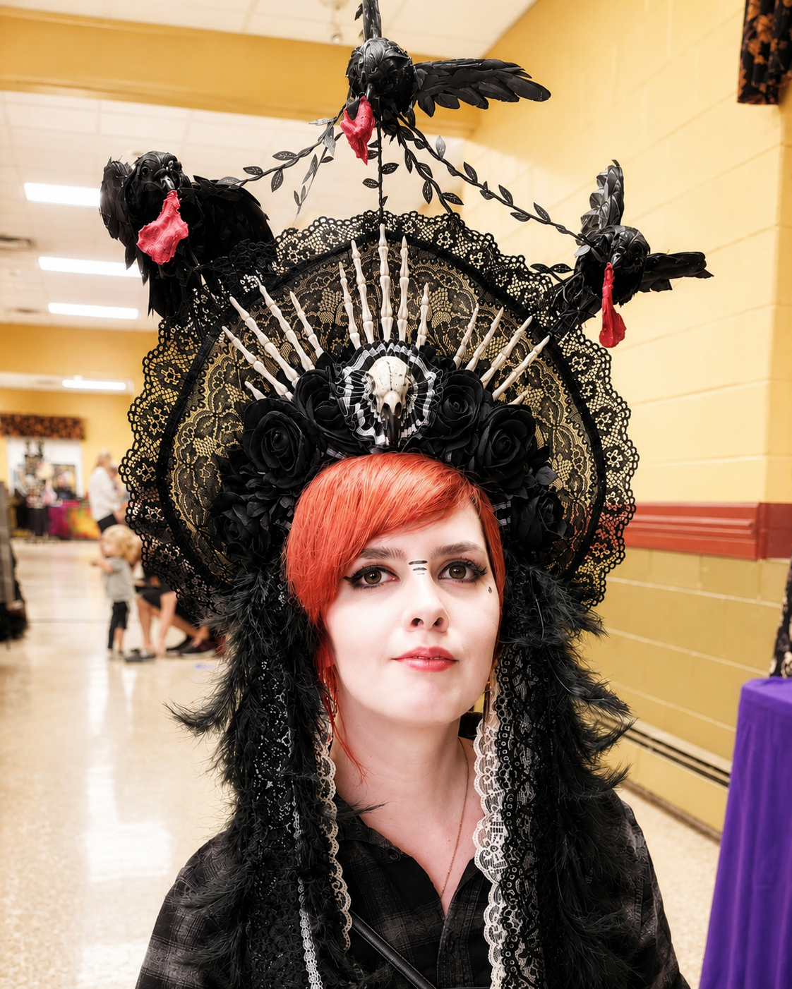 A woman with red hair wearing a black lace headdress decorated with black roses, a skull, and candles. The headdress has three black crow sculptures with red beaks puffing out red tissue paper. She has detailed makeup with tribal lines on her forehea