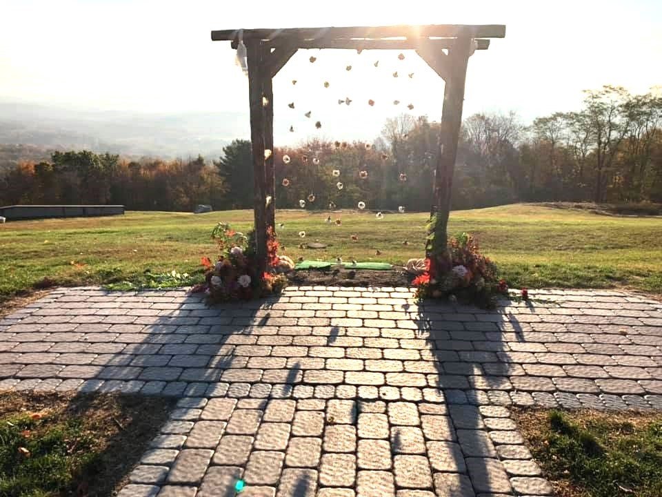Outdoor wedding arch decorated with flowers on a cobblestone path, with sunlight shining behind it and trees in the background.