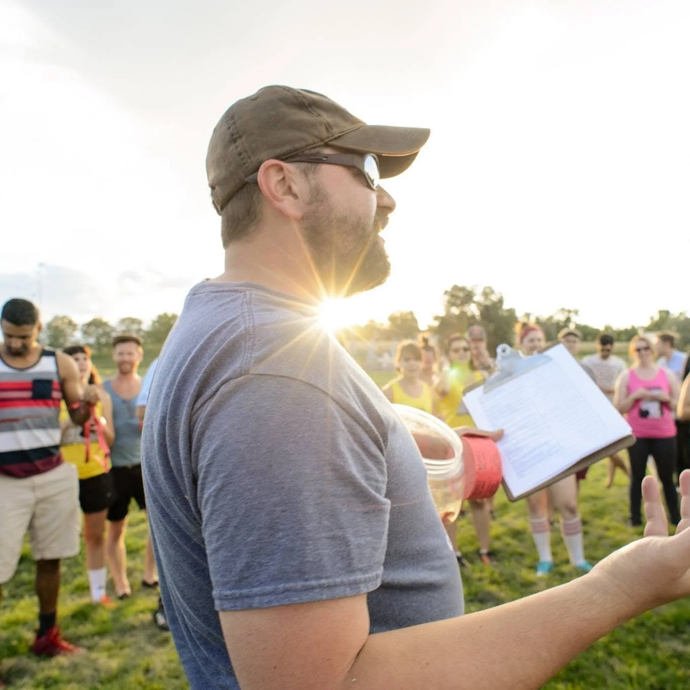 A man in a gray shirt, cap, and sunglasses reads from a paper while standing outdoors on a sports field during sunset. A group of people in colorful athletic attire stands in the background, smiling and listening.