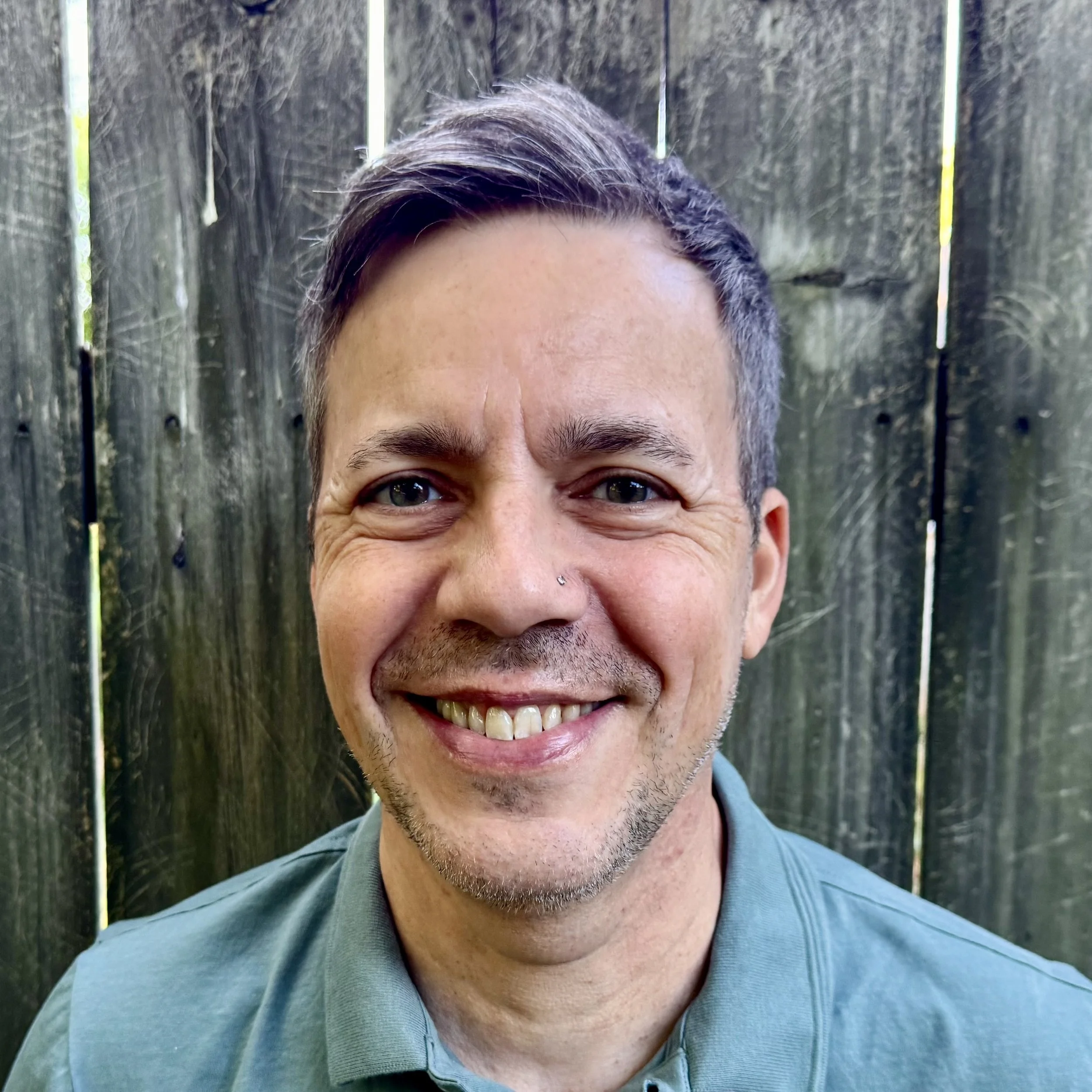 Close-up of a smiling man with short brown hair and a light beard, standing in front of a wooden fence.