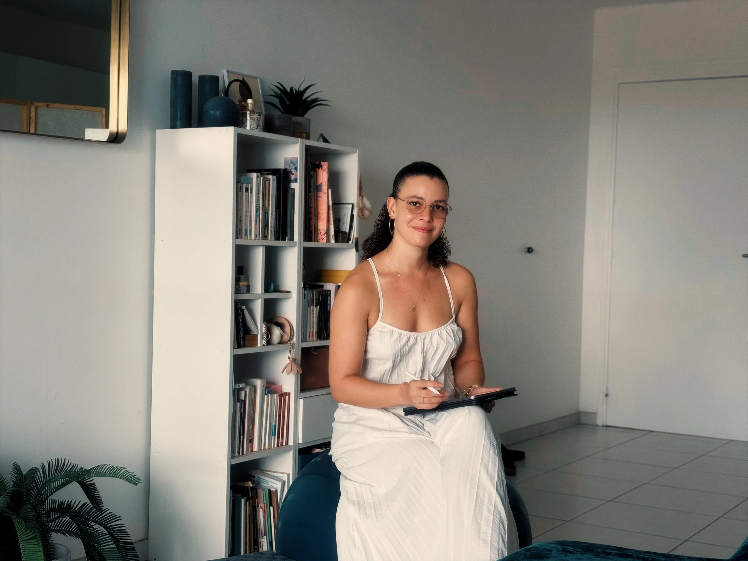 Une femme souriante portant des lunettes, assise dans un intérieur moderne près d'une bibliothèque blanche avec des livres, des objets décoratifs et une plante en pot, tenant un carnet ou un livre.