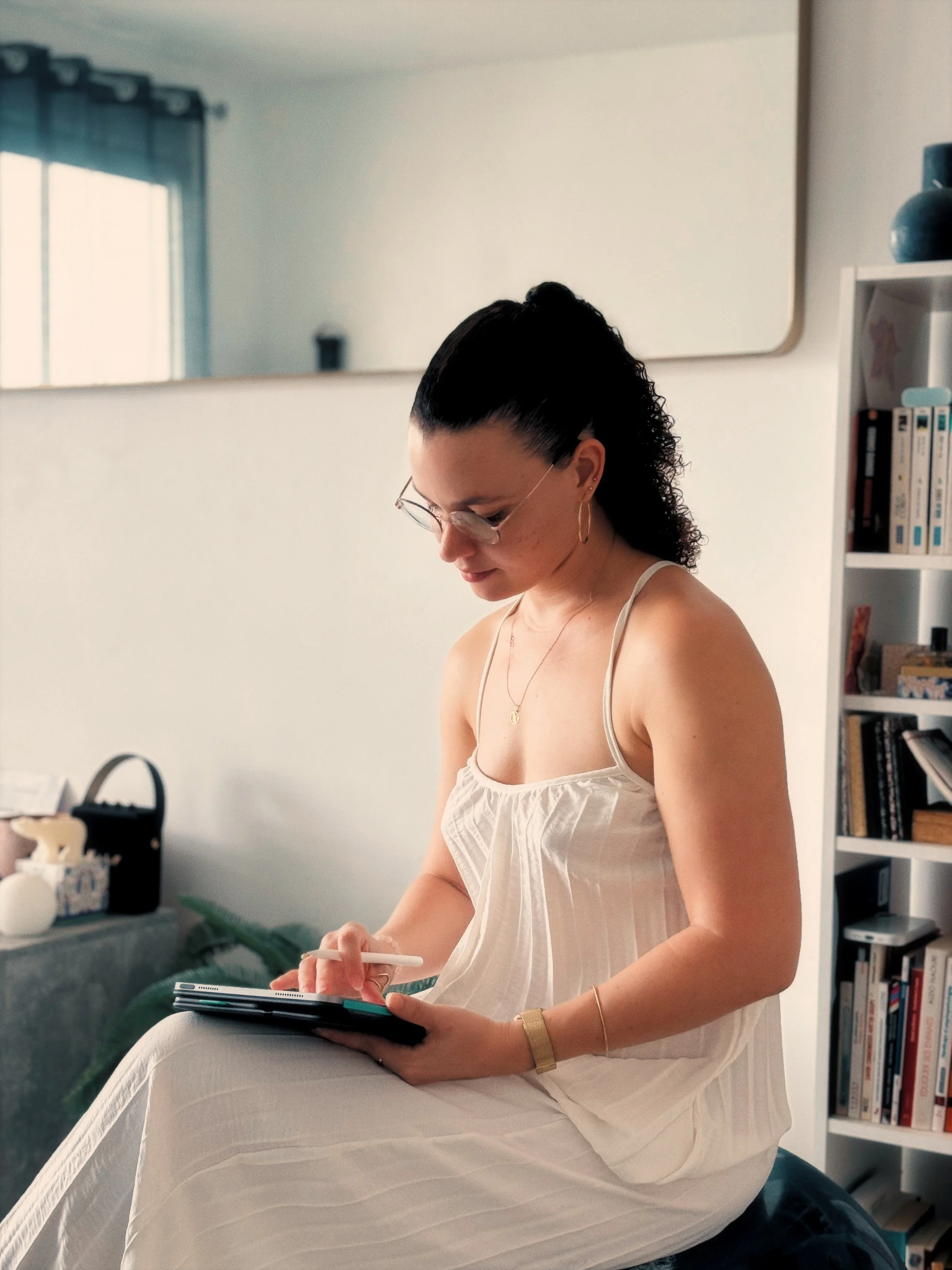 Une femme assise sur un canapé écrivant dans un carnet, portant une robe blanche, avec une bibliothèque à droite et une grande fenêtre à gauche dans une pièce lumineuse.