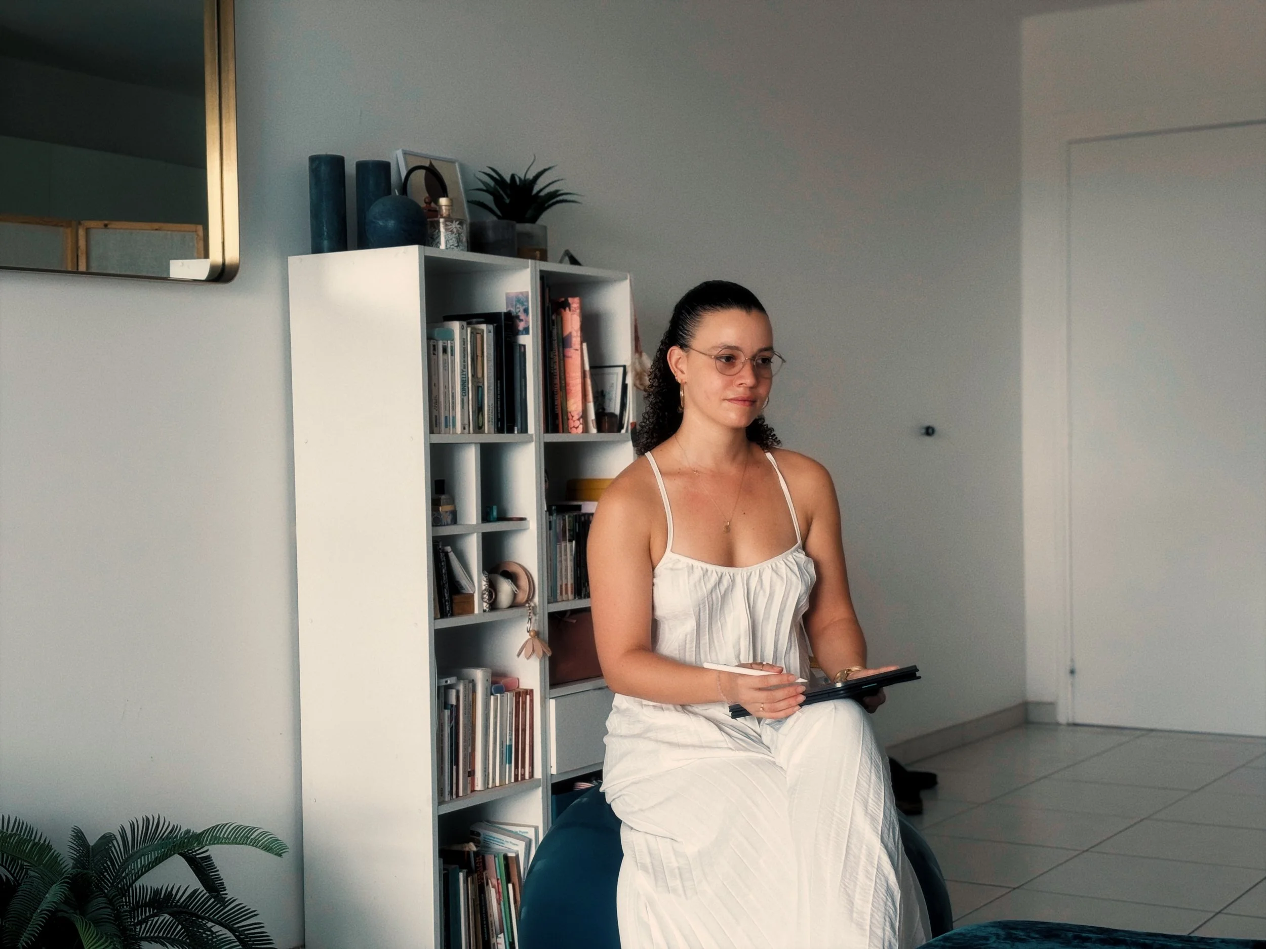 Jeune femme avec lunettes assise à côté d'une étagère remplie de livres, tenant une tablette, dans une pièce lumineuse avec une décoration moderne.