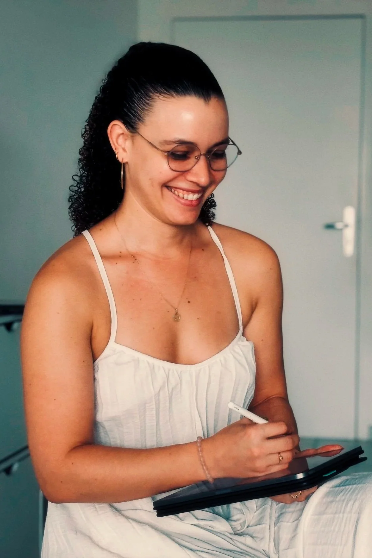 Une femme souriante avec des lunettes, portant un haut blanc à fines bretelles, utilisant une tablette tactile dans un intérieur lumineux.