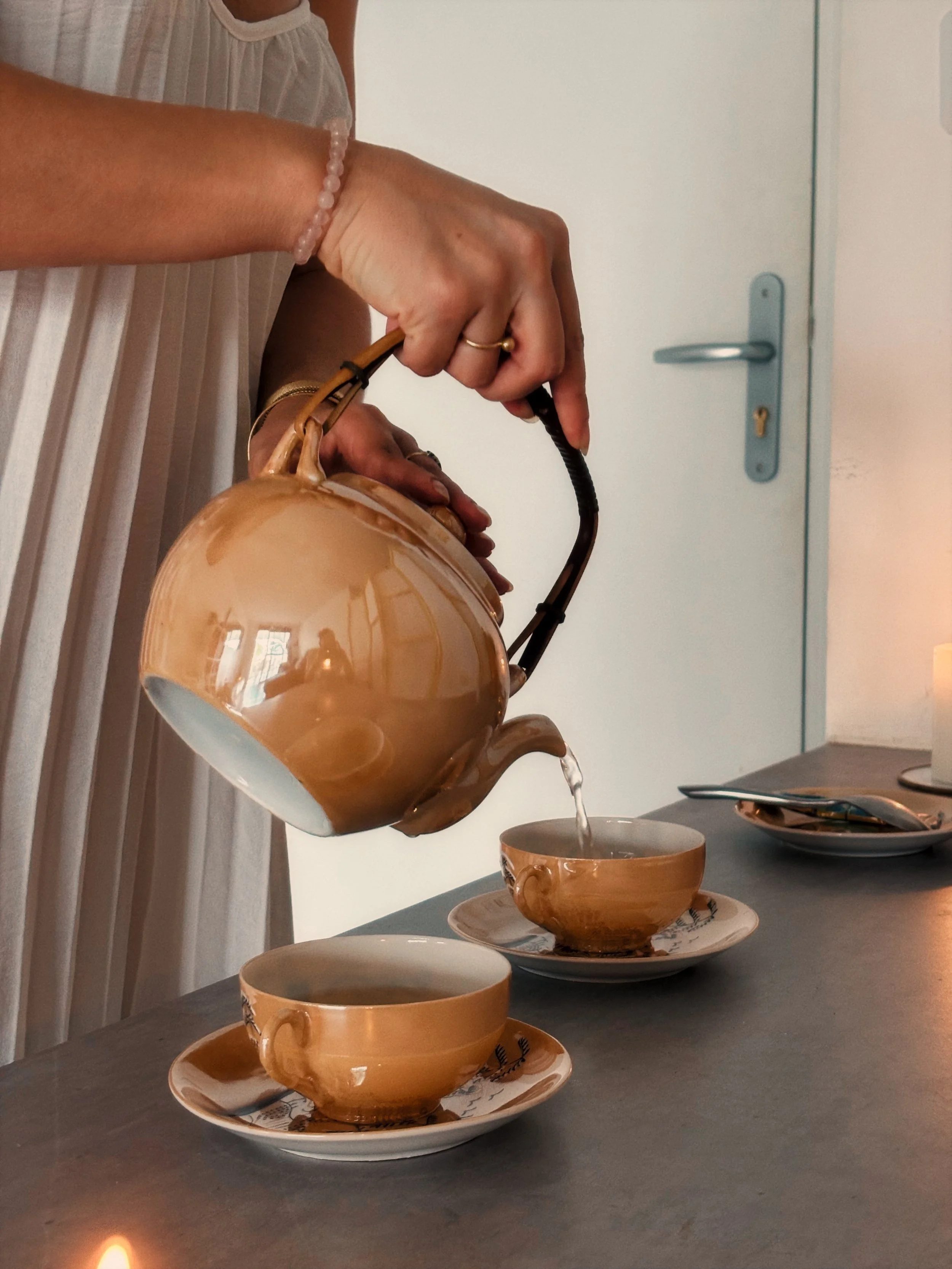 Une personne verse du café d'une théière en céramique dans une tasse sur une table. La tasse est placée sur une petite assiette. La scène se déroule dans une pièce avec une porte en arrière-plan.