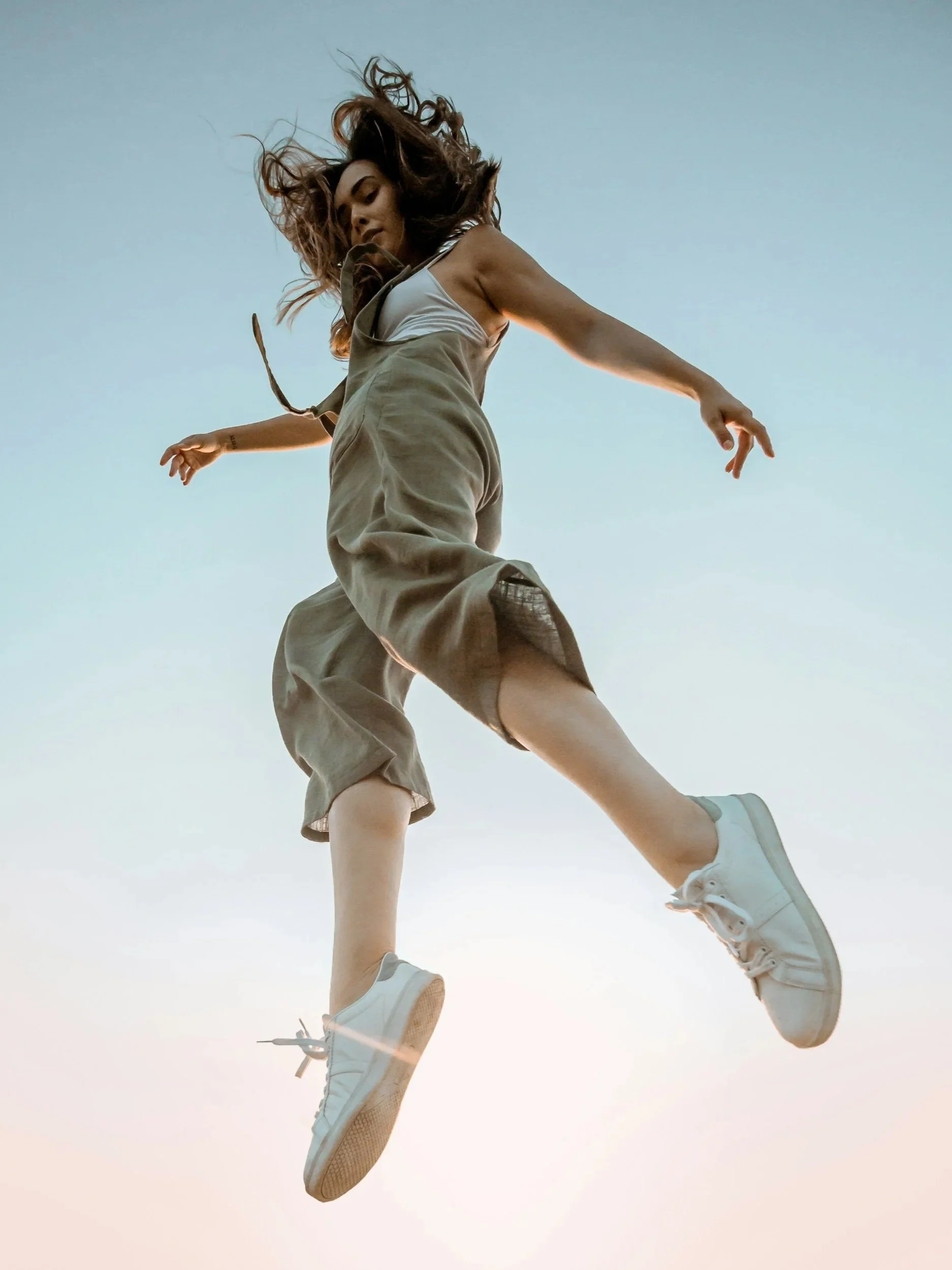A person wearing beige cargo shorts, a white tank top, white sneakers, and fishnet stockings, jumping against a clear sky during sunset.