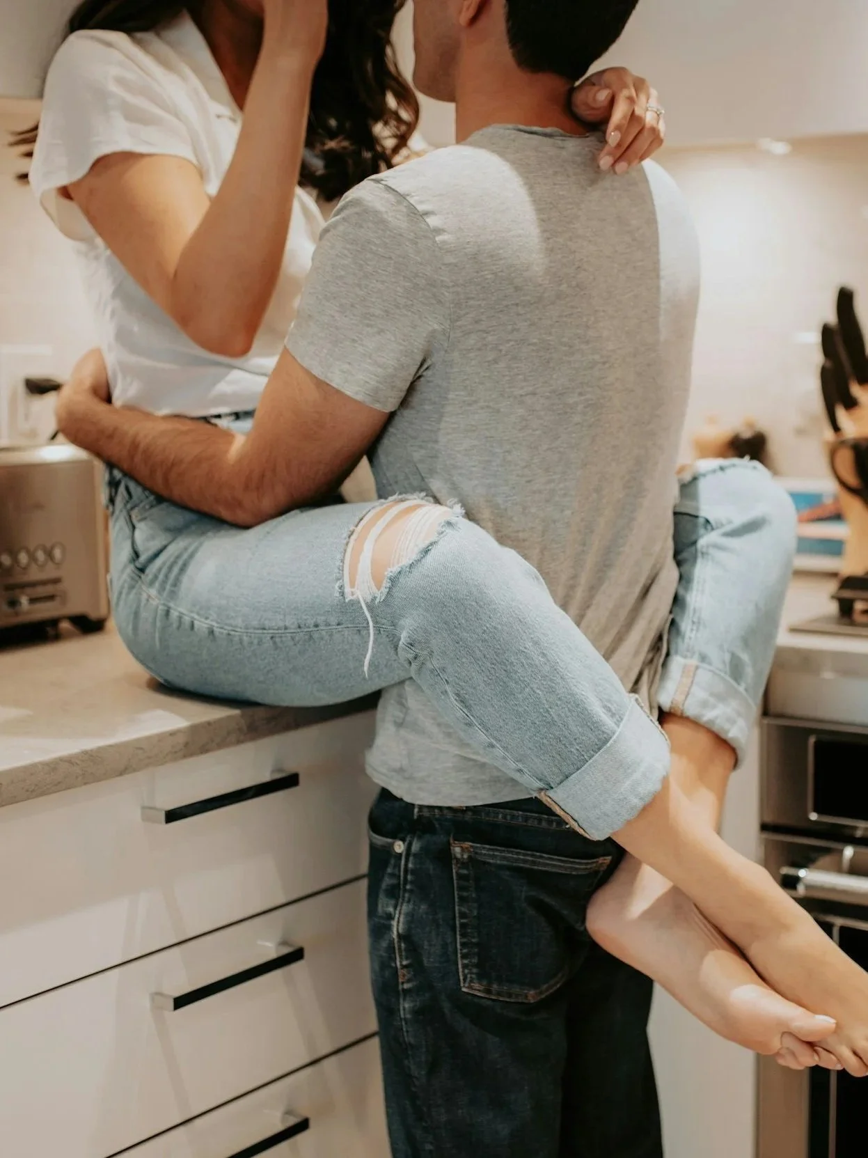 A couple kissing in a kitchen, with the woman sitting on a countertop and the man standing holding her with his shirt in the foreground.