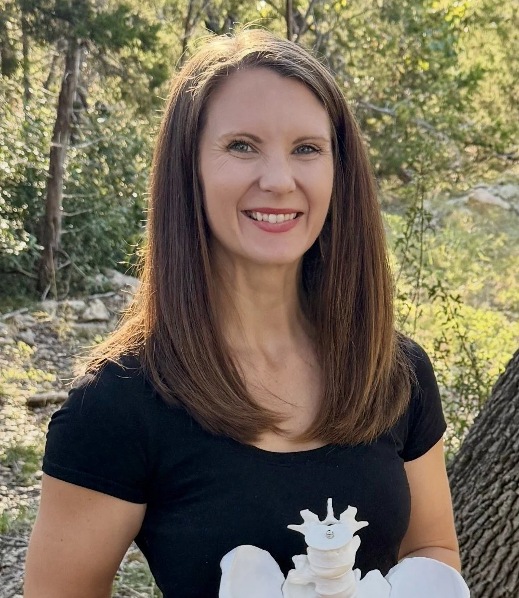 A woman with long brown hair smiling outdoors in natural sunlight, wearing a black t-shirt holding a model pelvis, standing in a wooded area.