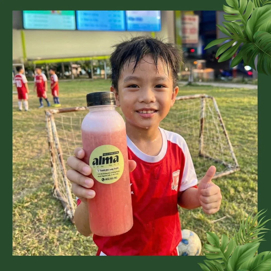 Young boy in a red soccer uniform holding a bottle of pink beverage, smiling, on a soccer field with goalposts and other children in the background.