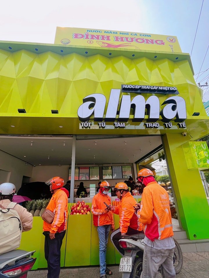 A street view of a green toko with a sign reading Alma and Vietnamese text, with delivery personnel in orange uniforms and helmets waiting outside.