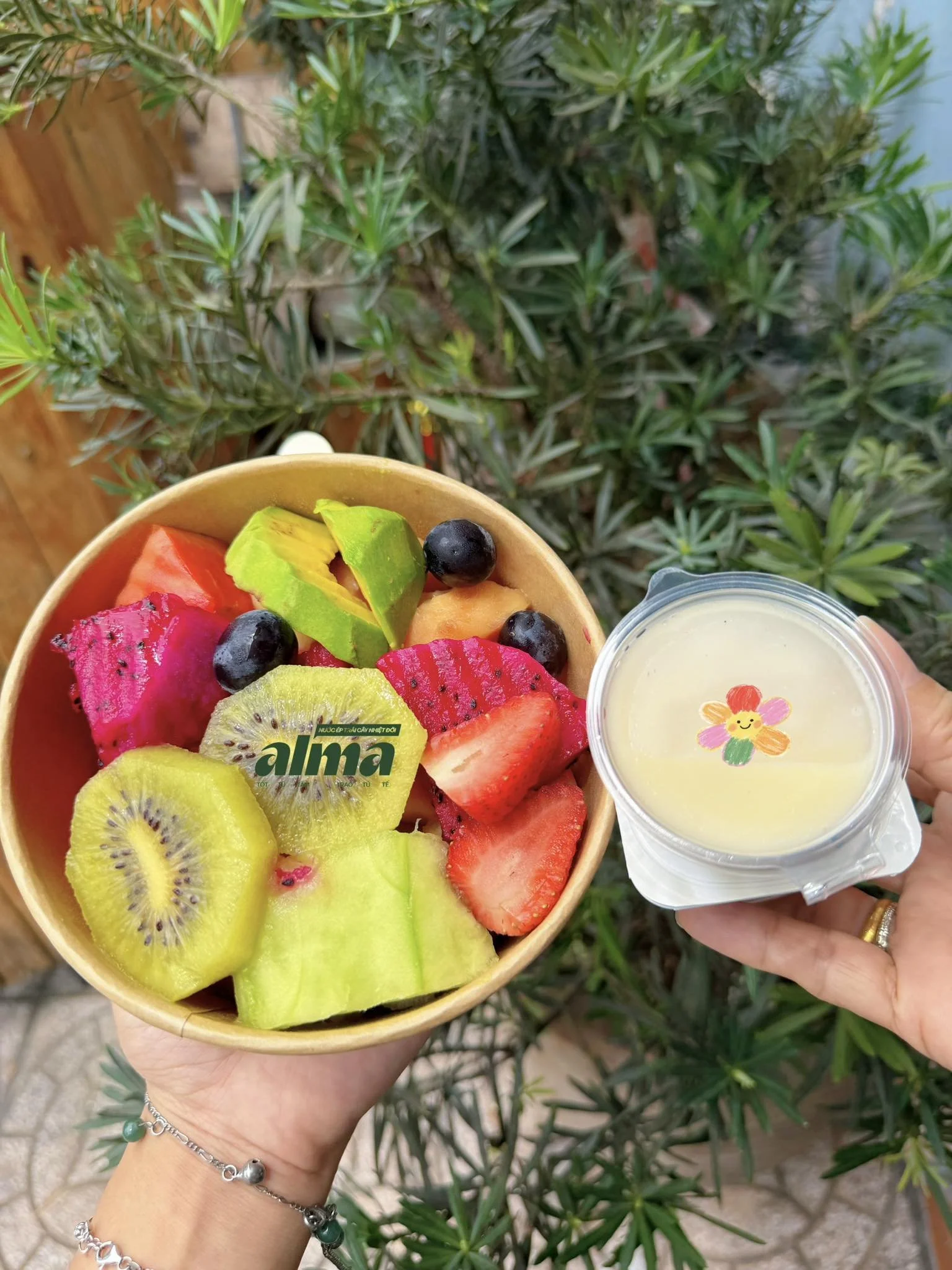 A person holding a wooden bowl filled with a variety of sliced fresh fruits including kiwi, dragon fruit, strawberries, and blueberries, along with a small cup of yellowish smoothie or juice, outdoors with green plants in the background.