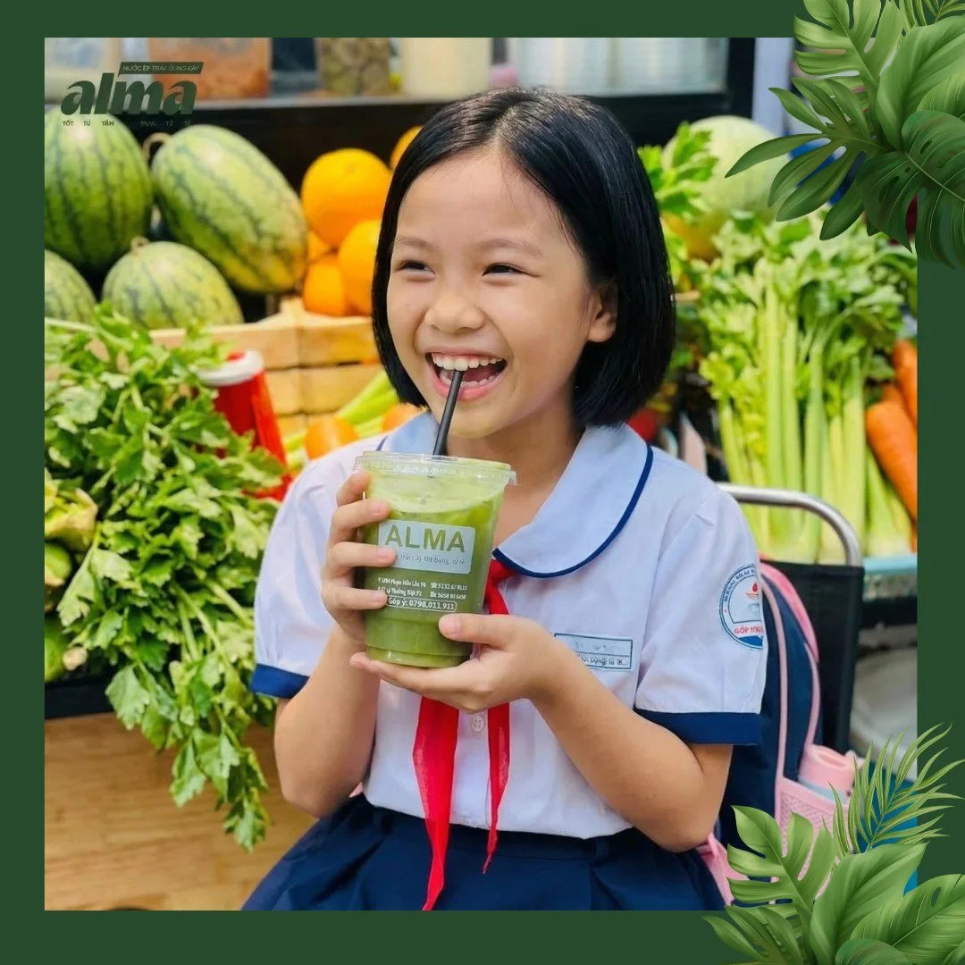 A young girl in a school uniform smiling and drinking a green smoothie at a produce market, surrounded by watermelons, oranges, carrots, and leafy greens.