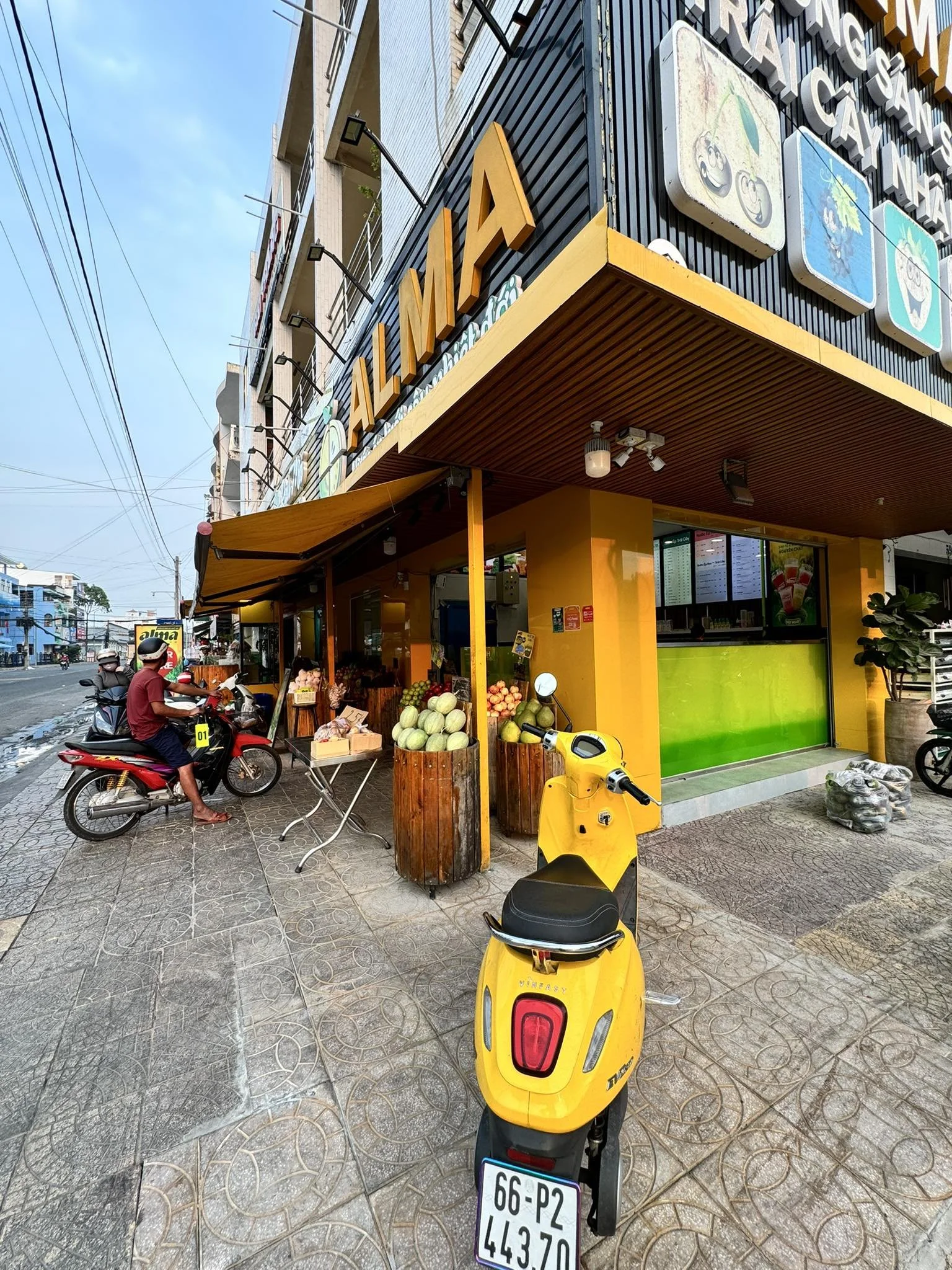 A street corner with a small fruit stand selling melons and other produce, a man riding a motorcycle, and a yellow scooter parked in front. The building behind has a sign reading 'ALMACA' in large yellow letters, with a store selling beverages on the ground floor. The sky is blue with some clouds.