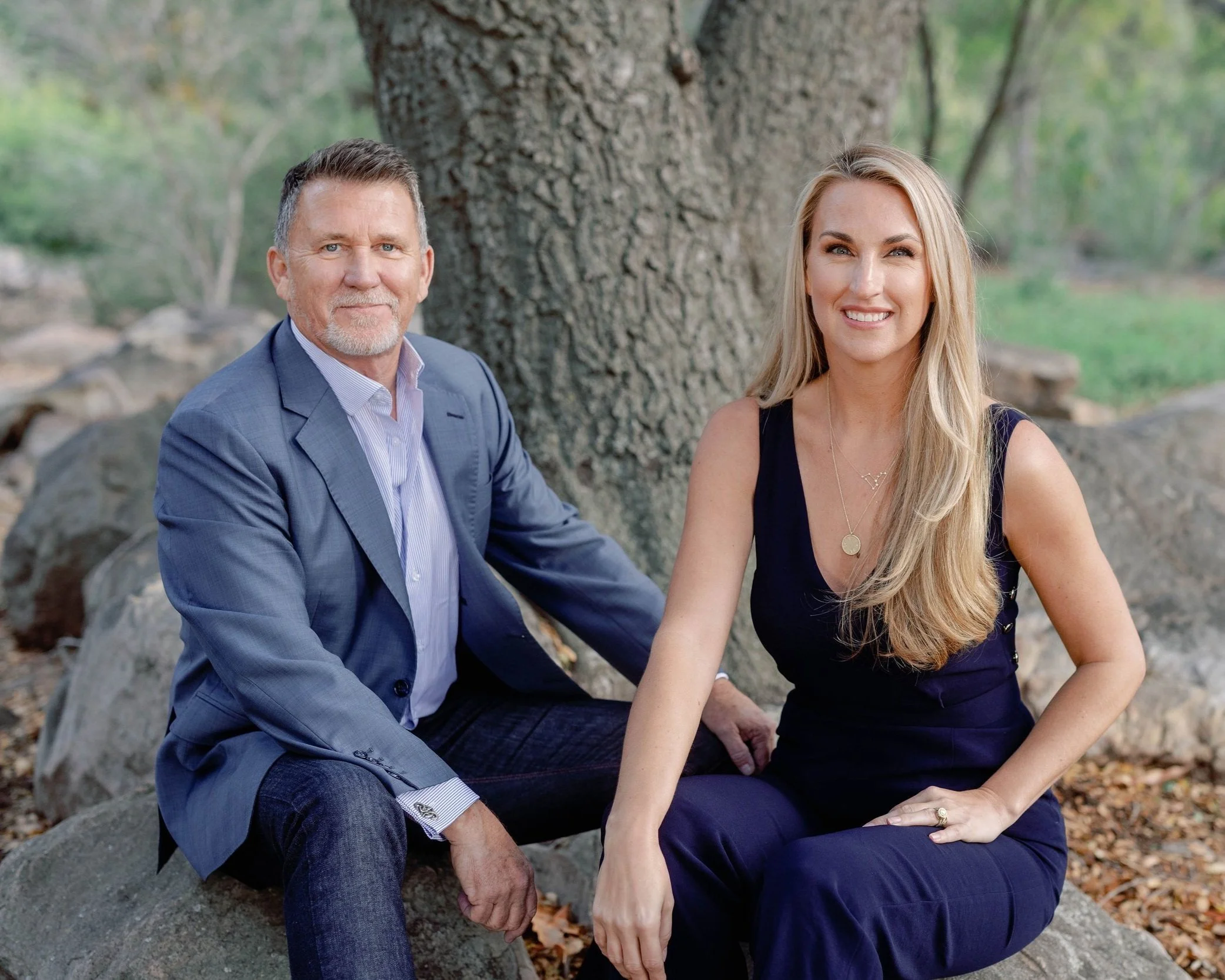 A middle-aged man in a blue suit and a young woman in a dark blue dress sitting outdoors near a tree.
