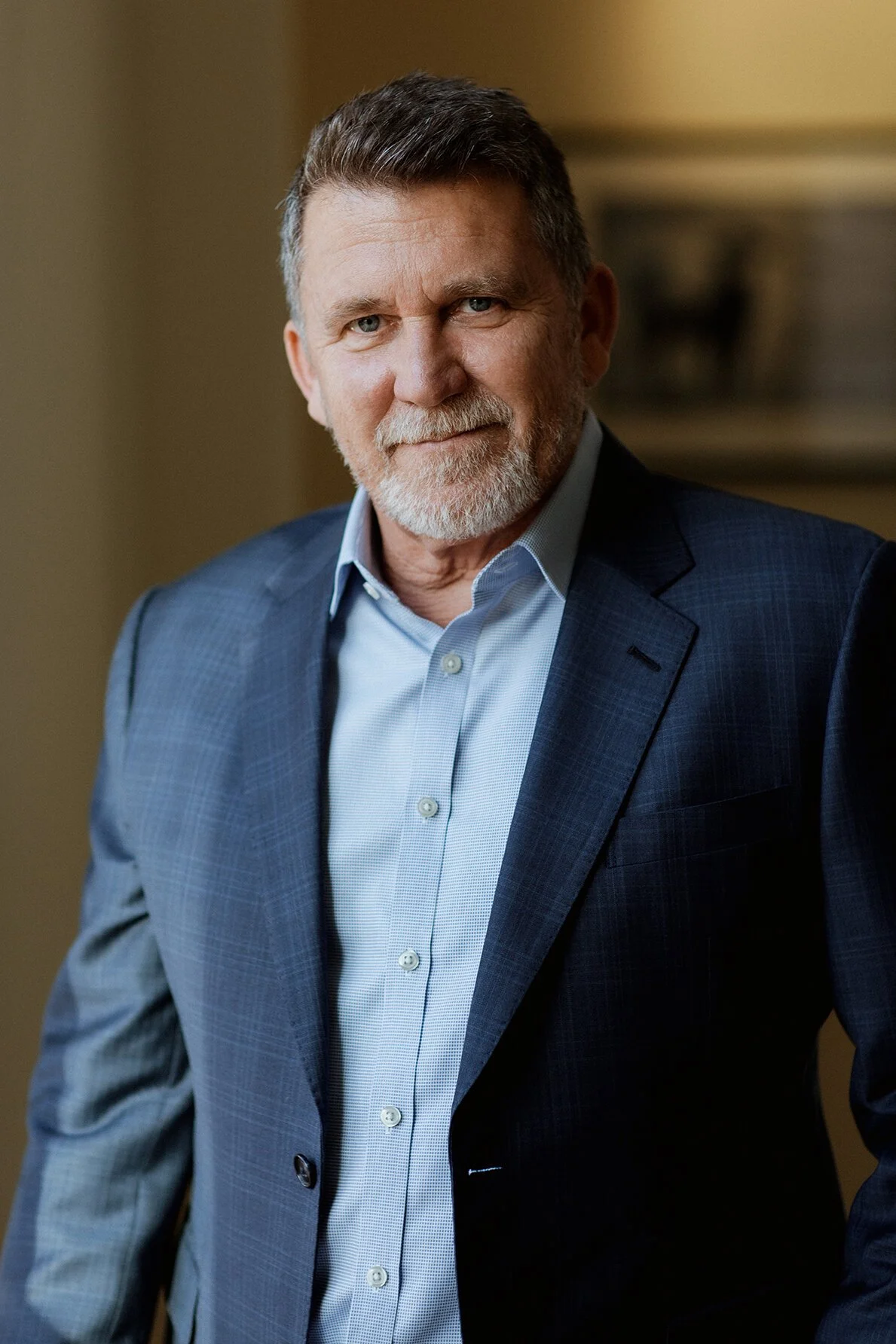 A middle-aged man with gray hair and a beard, wearing a blue suit jacket and a light blue dress shirt, standing indoors with a neutral background.