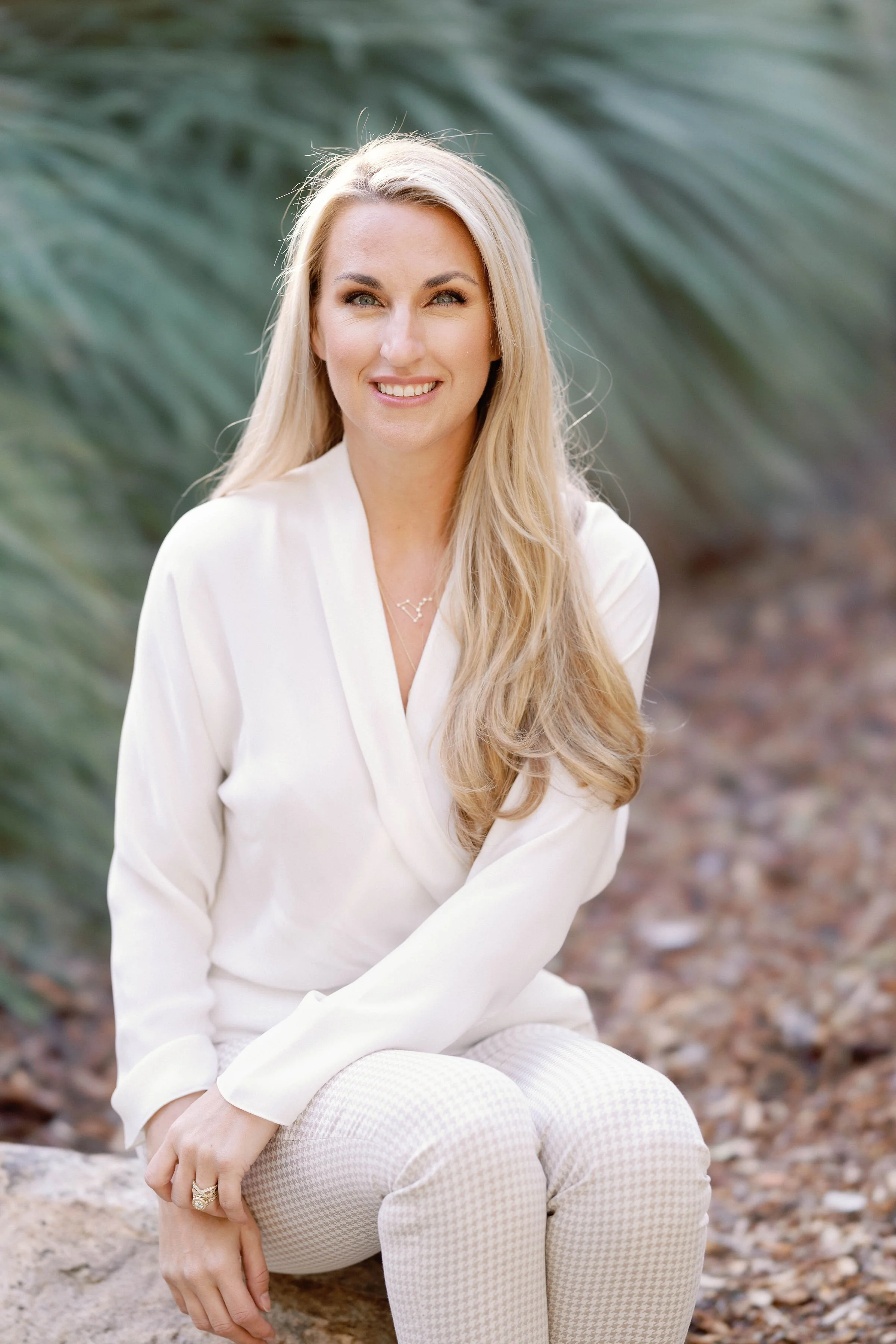 A woman with long blonde hair, smiling, wearing a white blouse and light-colored pants, sitting on a rock outdoors with a blurred natural background.
