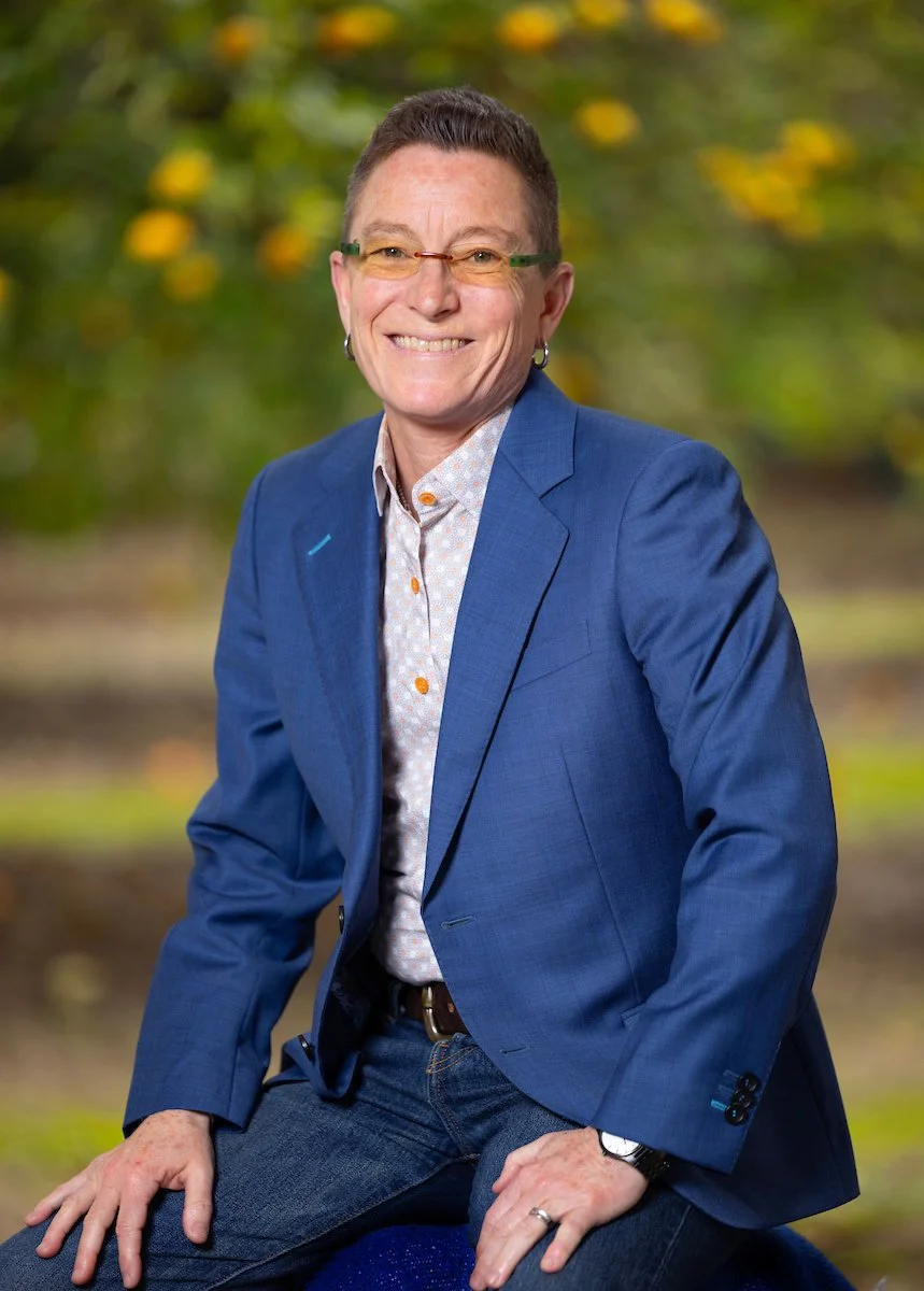 A woman with short hair and glasses, wearing a blue blazer and a patterned shirt, sitting outdoors with a blurred background of trees with yellow and green leaves, smiling at the camera.