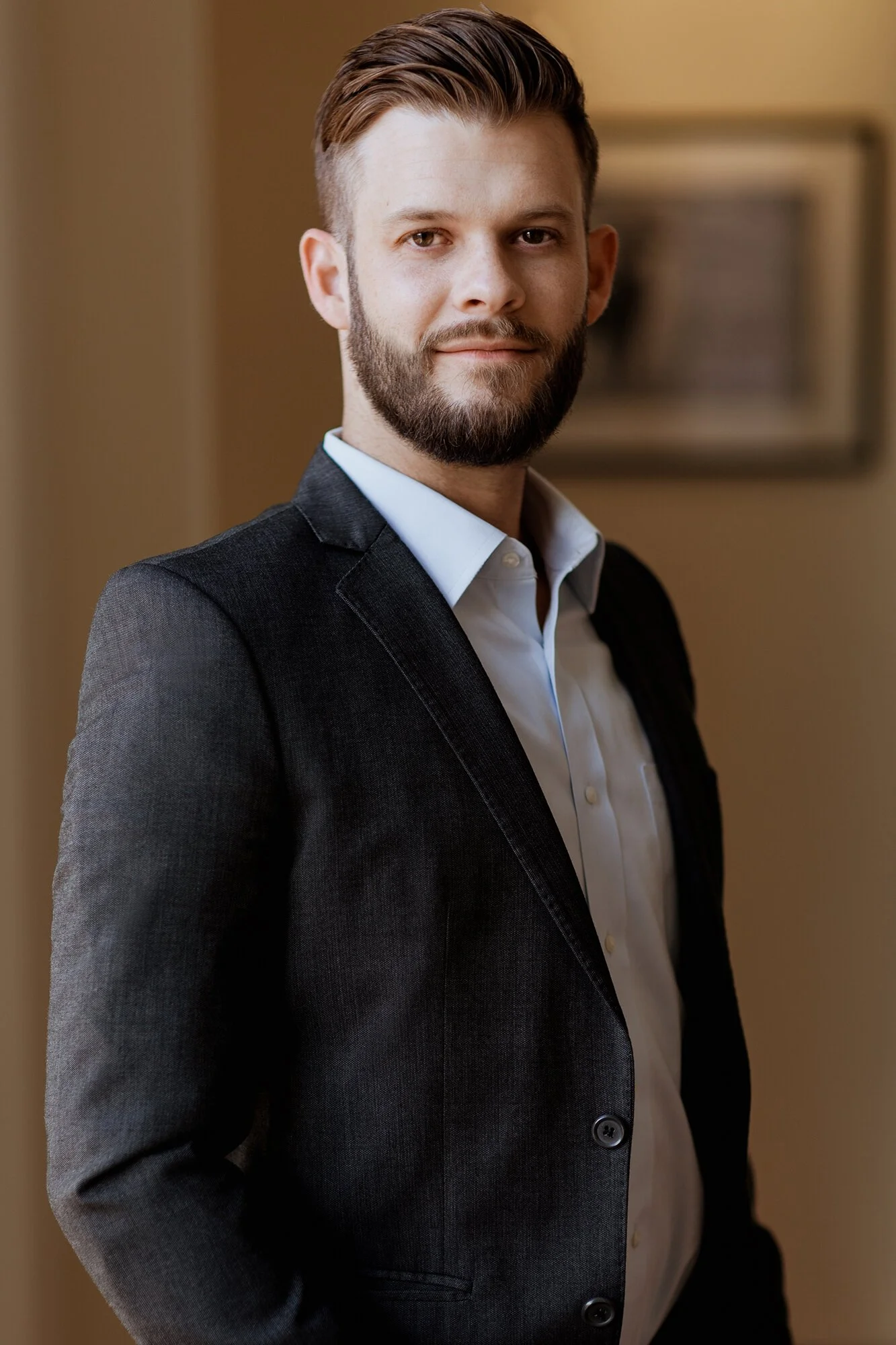 A young man with a beard and short, styled hair, dressed in a dark suit and white shirt, standing indoors with a warm-toned background