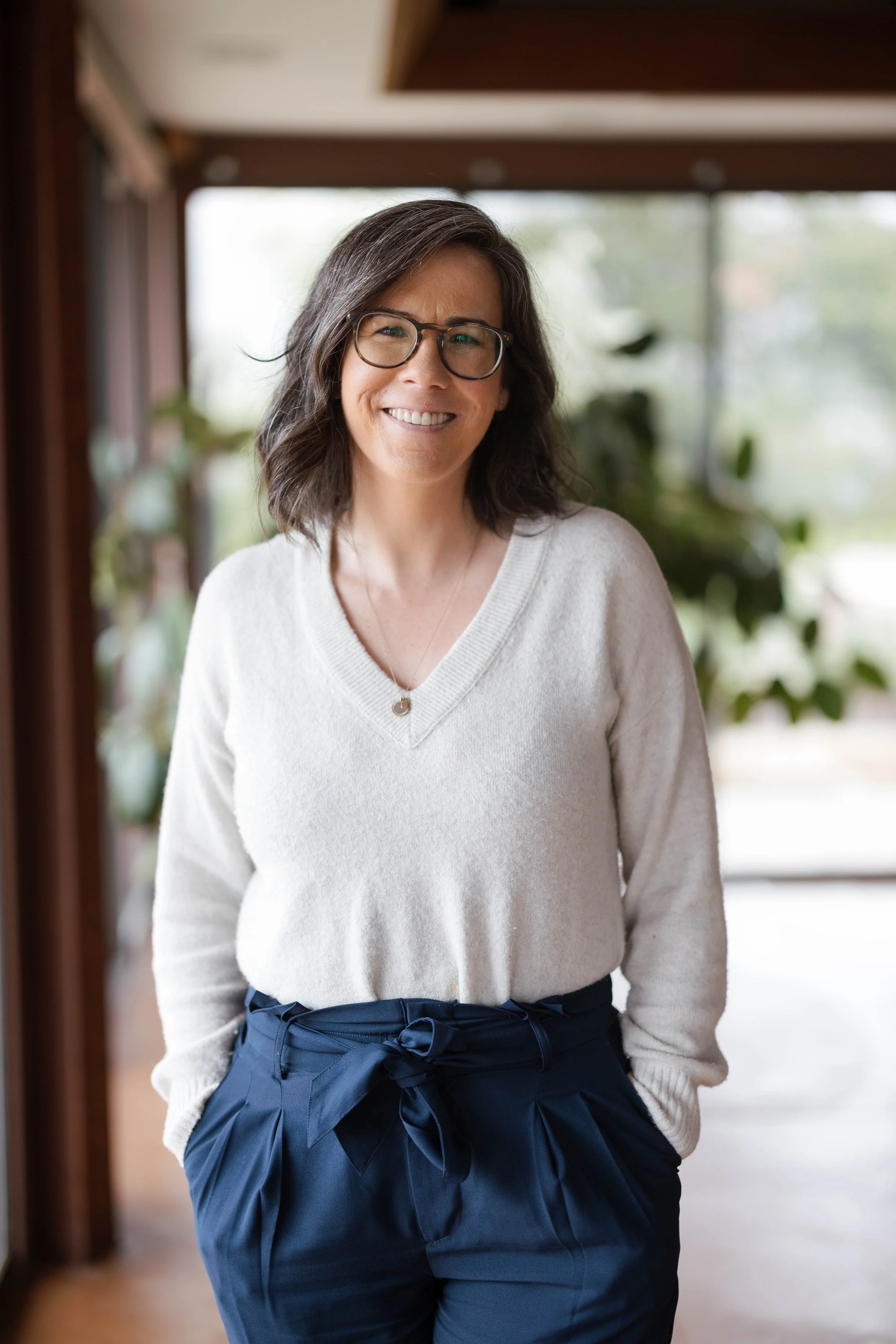 A woman with shoulder-length wavy dark hair, glasses, and a light smile standing indoors near a large window with blurred greenery outside. She is wearing a white V-neck sweater and navy blue pants with a tied waist.